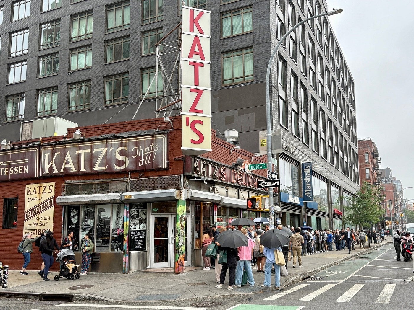 A long line of people with umbrellas waits outside Katz's Delicatessen—a classic among road trip food stops—on a city street corner, with tall buildings and storefronts visible in the background on a cloudy day.