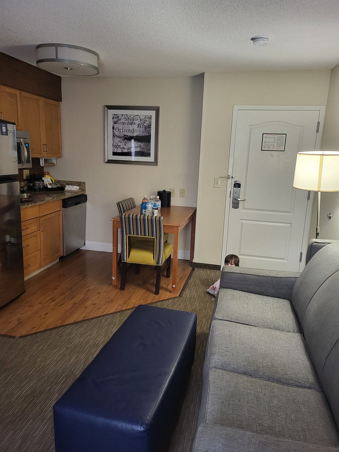 A hotel room from one of the top Hotel Brands 2026, featuring a small kitchen, dining table with chairs, and a gray sofa. Near the door, a young child is partially visible, peeking from behind the wall. Bottled water and snacks sit on the table.