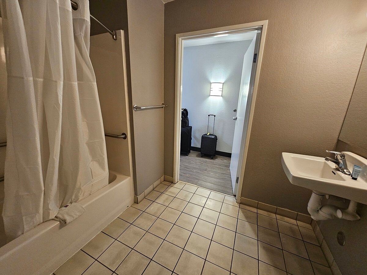 Hotel bathroom with tiled floor, a shower with curtain and grab bar, a wall-mounted sink stocked with toiletries—reflecting the thoughtful touches expected from leading Hotel Brands 2026. An open door reveals a hallway, suitcase, and lit wall lamp.