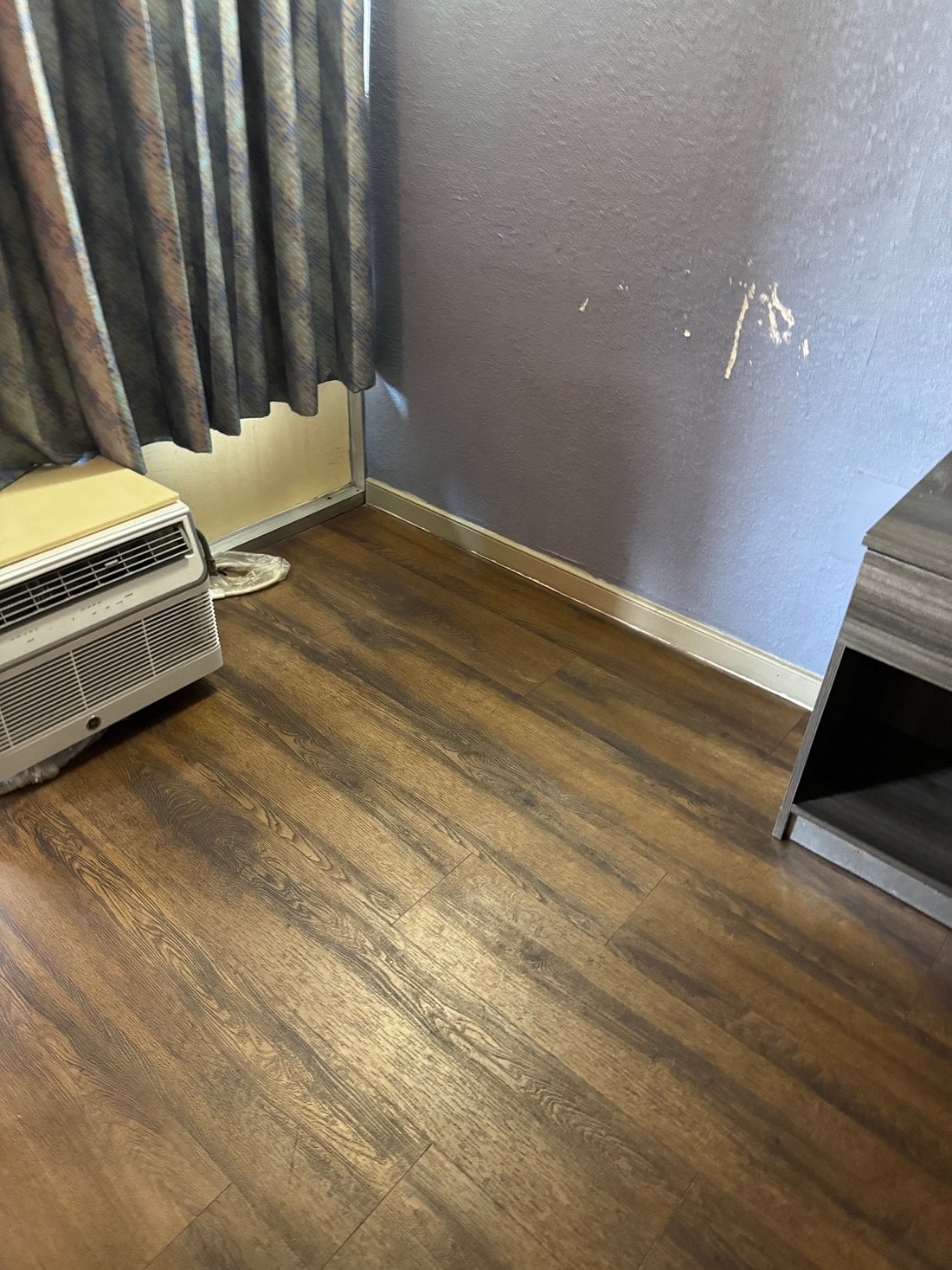 A corner of a room with wood-style flooring, a vintage air conditioning unit, dark-patterned curtains, a scuffed purple wall with peeling paint, and part of a wooden nightstand—an unexpected scene among future Hotel Brands 2026.