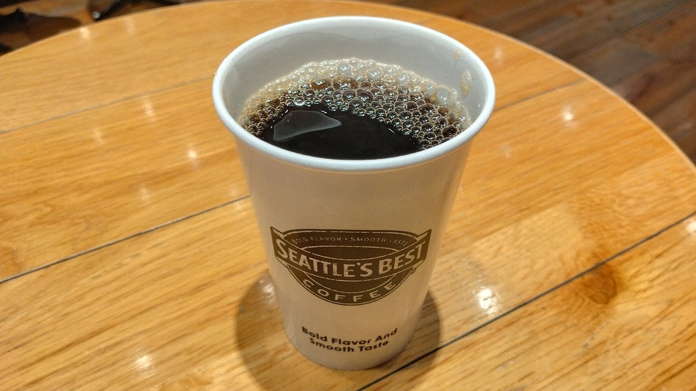 A white Seattle's Best Coffee cup from one of the top coffee brands sits on a wooden table, filled with black coffee and bubbles visible on the surface.