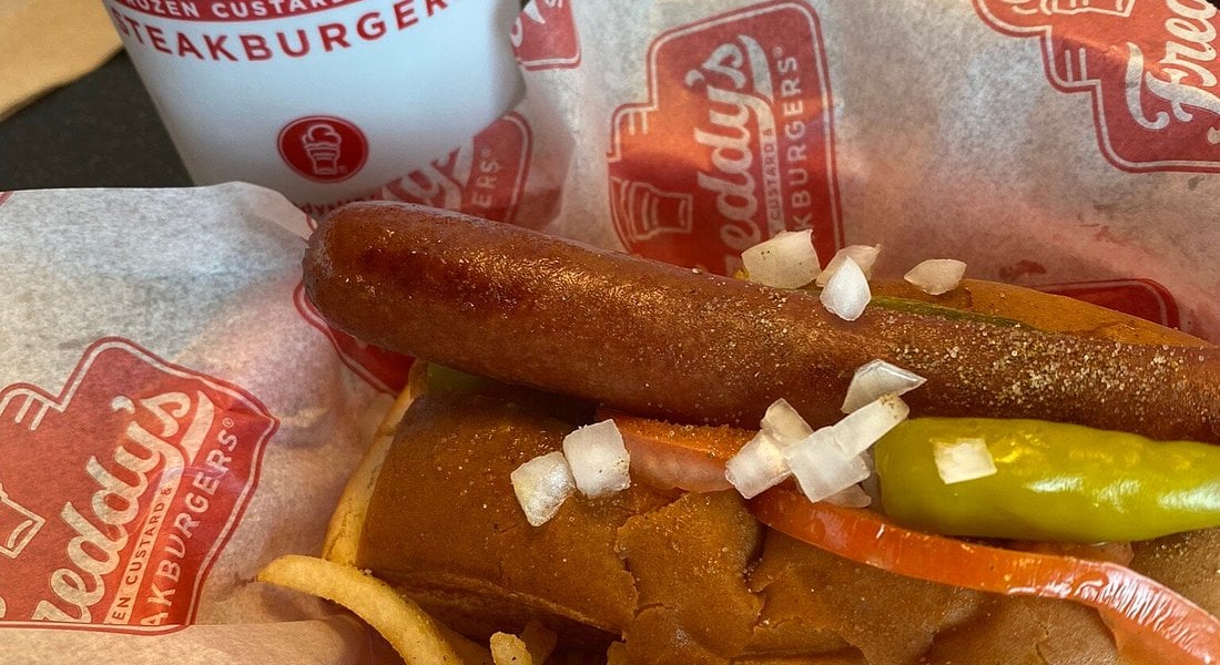 A Fast-Food Hot Dog topped with chopped onions, a pickle spear, and tomato on a bun, served on Freddy's branded paper next to a cup featuring Freddy's logo.