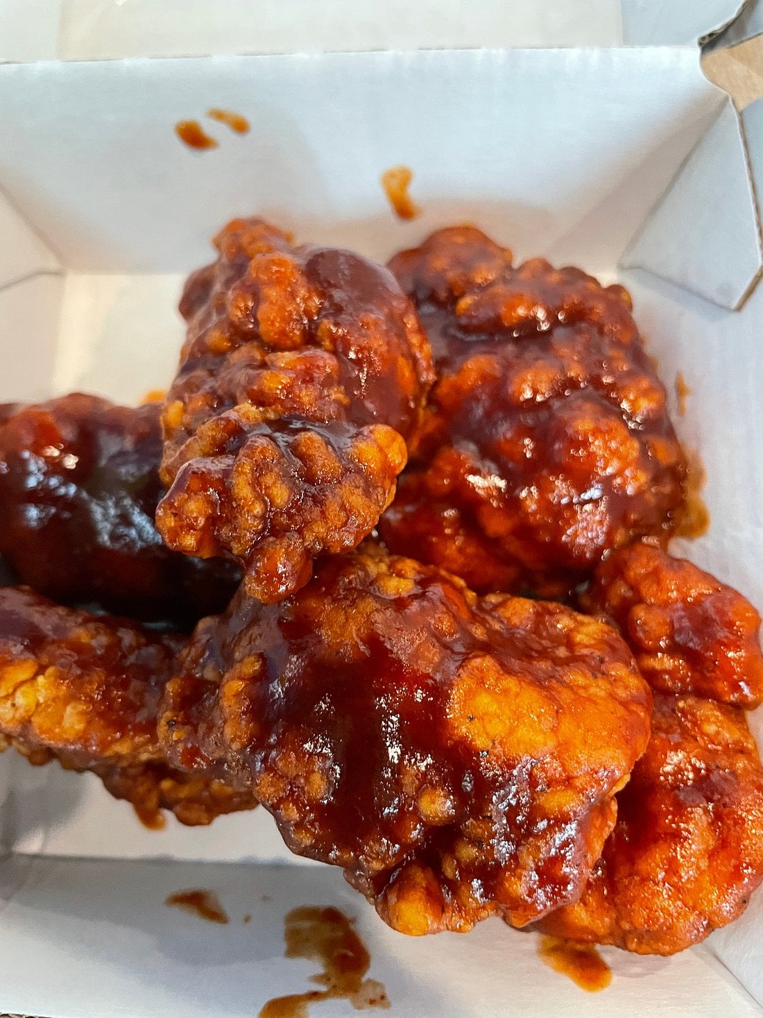 A close-up of several crispy fried chicken wings coated in a thick, glossy barbecue sauce, placed inside a white takeout box.