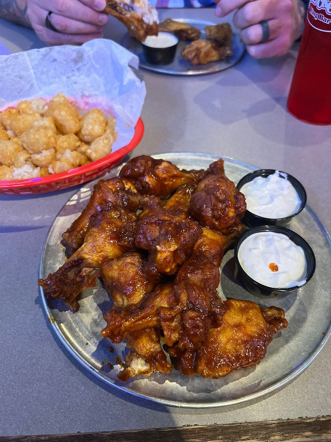 A plate of saucy buffalo chicken wings with two cups of ranch dressing sits on a table next to a basket of tater tots. Two people’s hands are seen in the background, holding food and dipping into sauce.