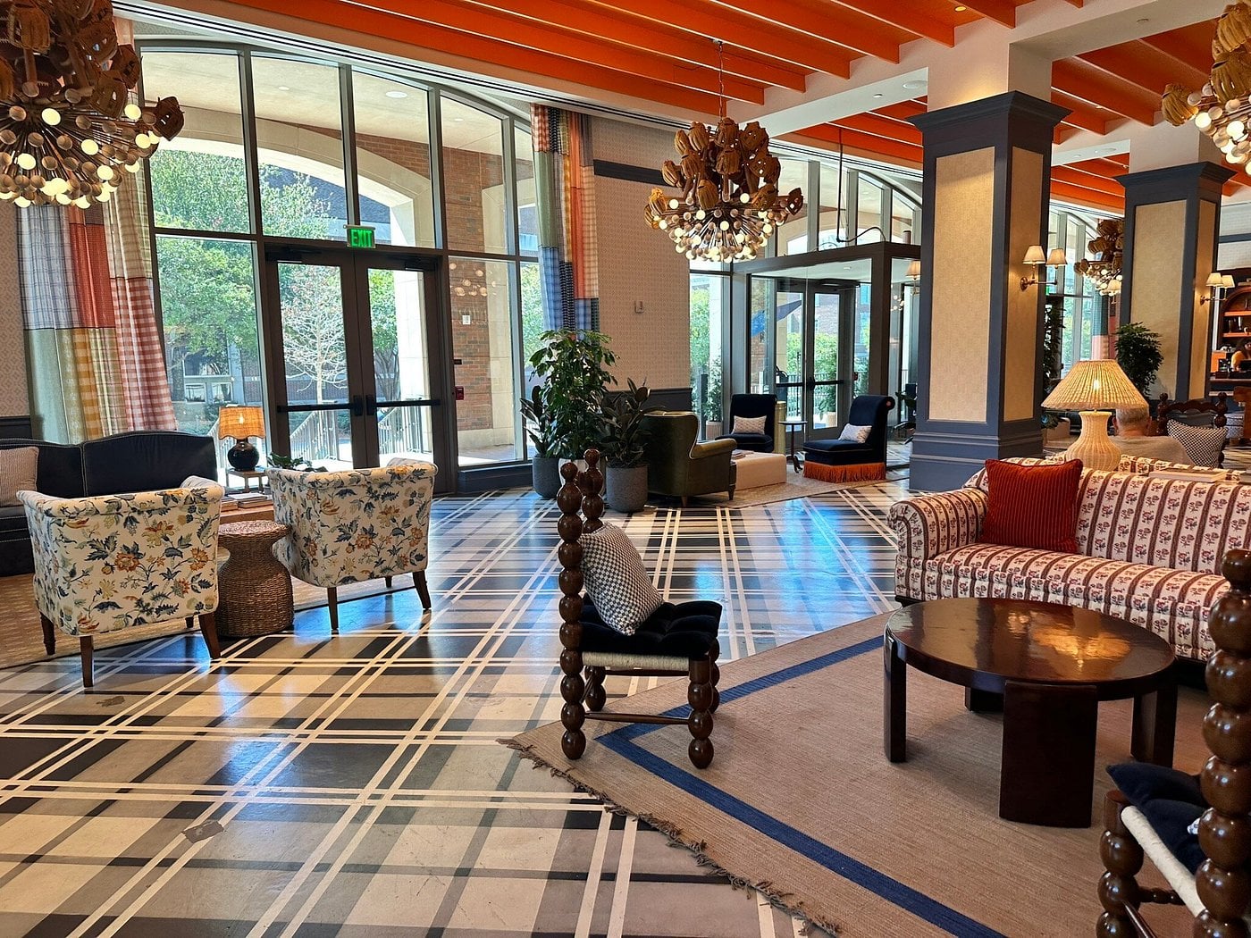 The lobby of this Small-Town Luxury Hotel features patterned armchairs, a striped sofa, round wooden tables, unique chandeliers, large windows with natural light, striking orange ceiling beams, and a black and white geometric floor design.