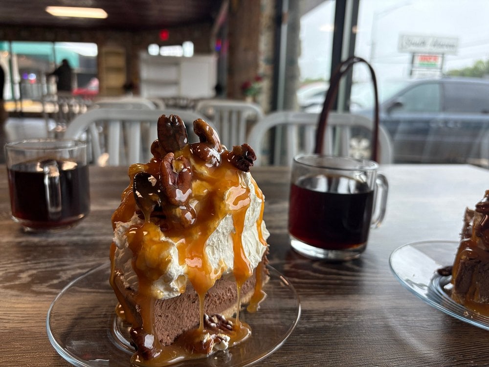 A slice of chocolate cake topped with whipped cream, caramel sauce, and pecans sits on a plate next to two cups of black coffee on a wooden table in a cafe with large windows and white chairs.