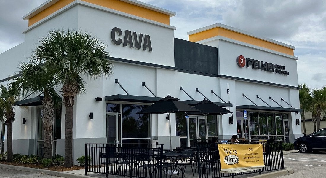 A modern restaurant building with signs for "CAVA" and "Pei Wei Asian Kitchen." There is a small outdoor seating area with black tables and umbrellas, and a yellow banner reading "We're Hiring" is displayed on the fence. Palm trees are nearby.