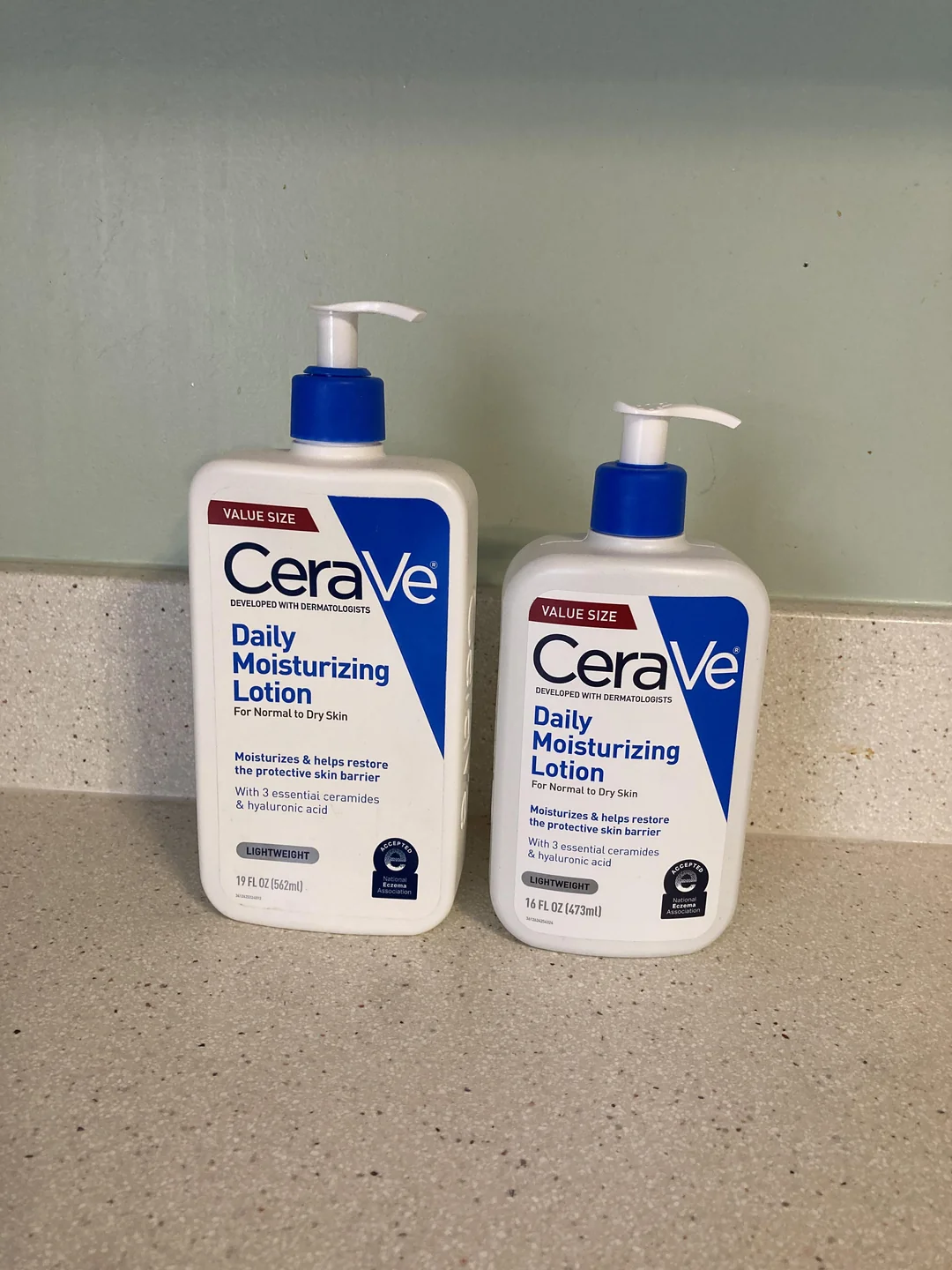 Two value size bottles of CeraVe Daily Moisturizing Lotion sit on a speckled countertop against a pale green wall, one with a pump (19 fl oz) and one without (16 fl oz).