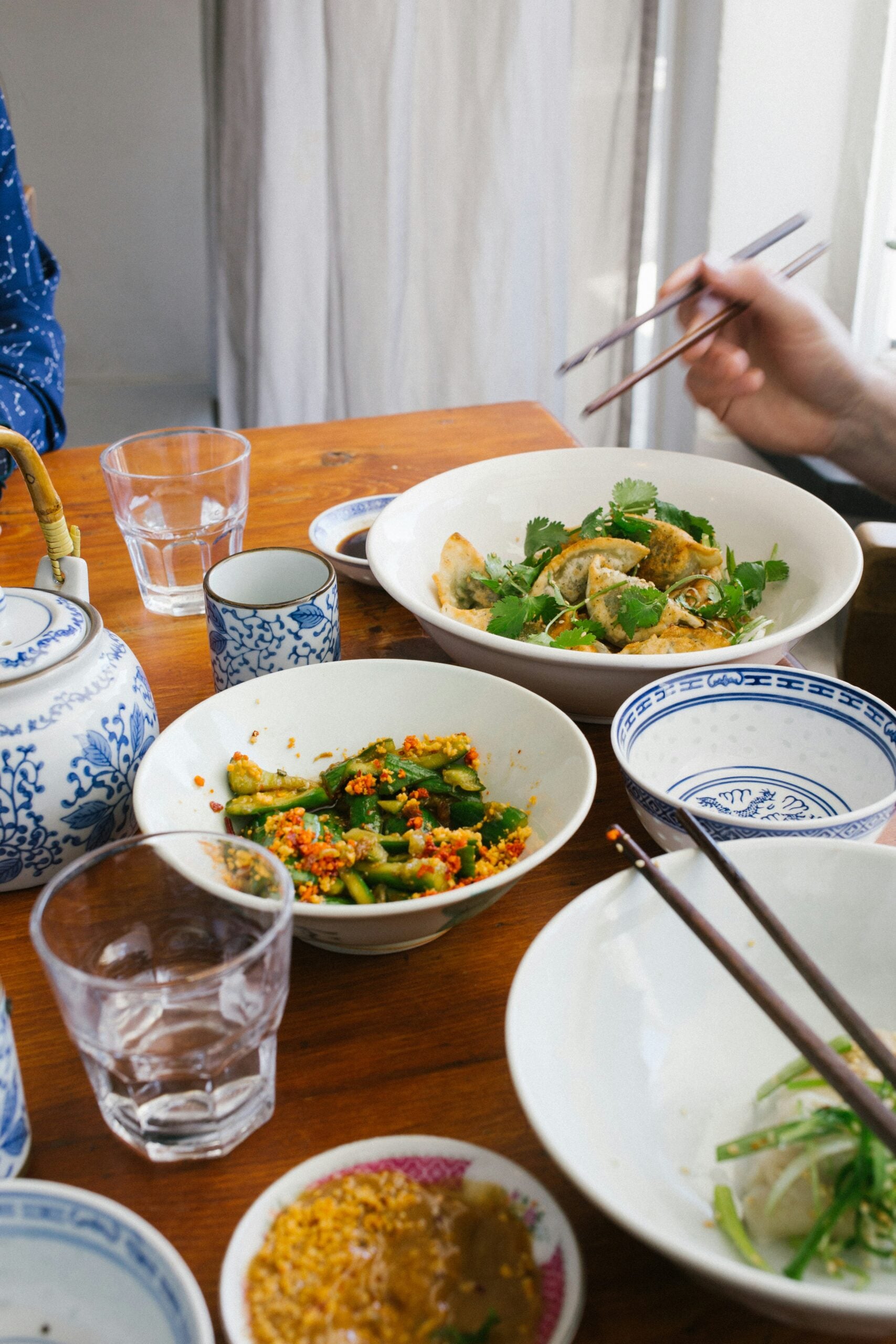 A wooden table set with blue and white dishes, cups, a teapot, and various Asian-inspired dishes, including noodles and vegetables. Two people are using chopsticks, and there are glasses of water on the table.
