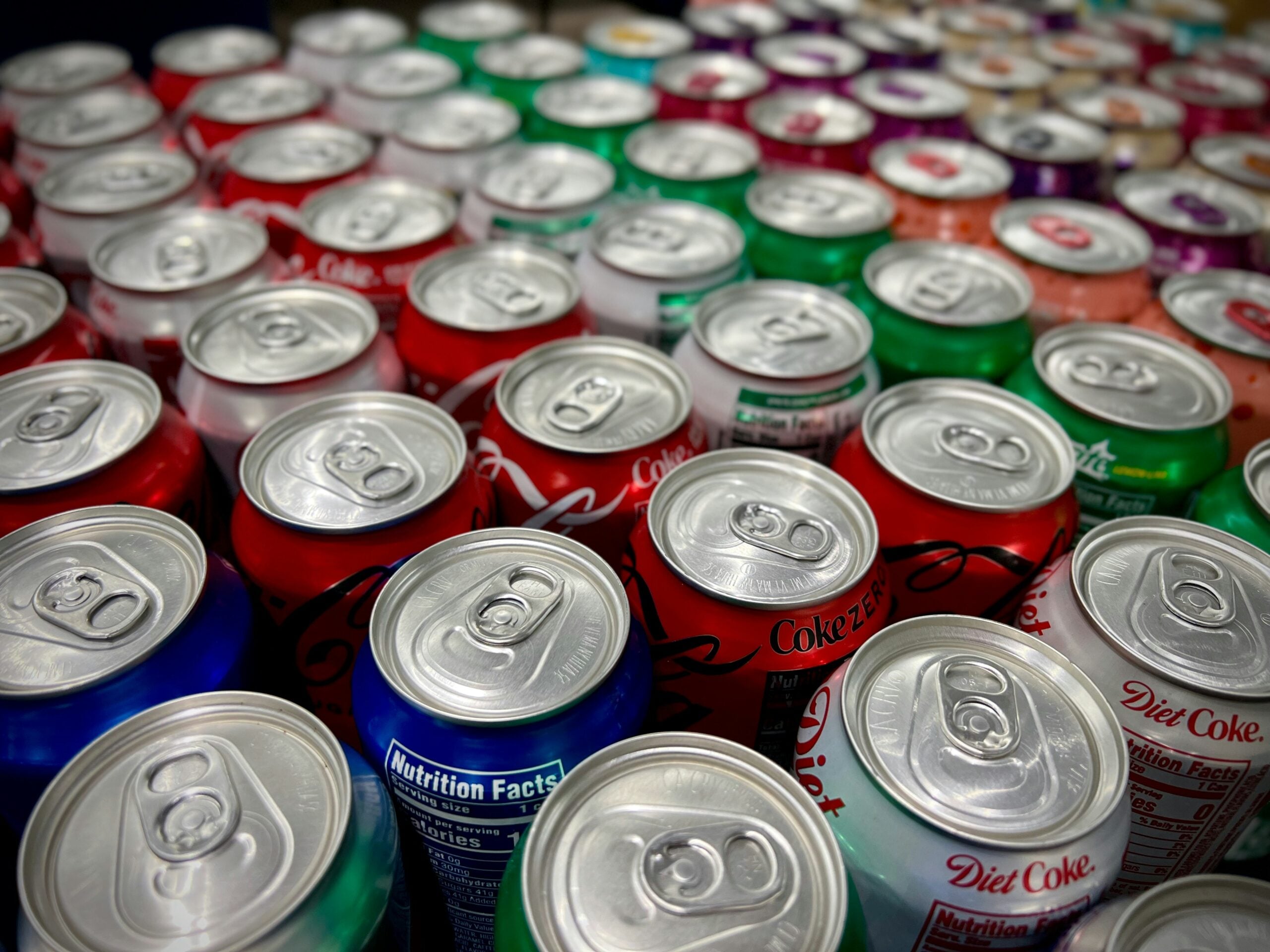A close-up view of many colorful soda cans, including red, green, blue, and silver varieties, arranged in rows with the tops and pull tabs visible.
