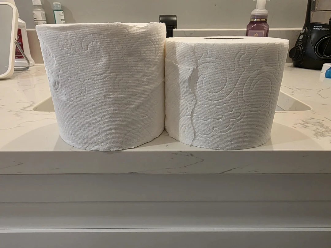 Two toilet paper rolls are side by side on a bathroom counter, with the roll on the left noticeably larger than the roll on the right. Bathroom items are visible in the background.
