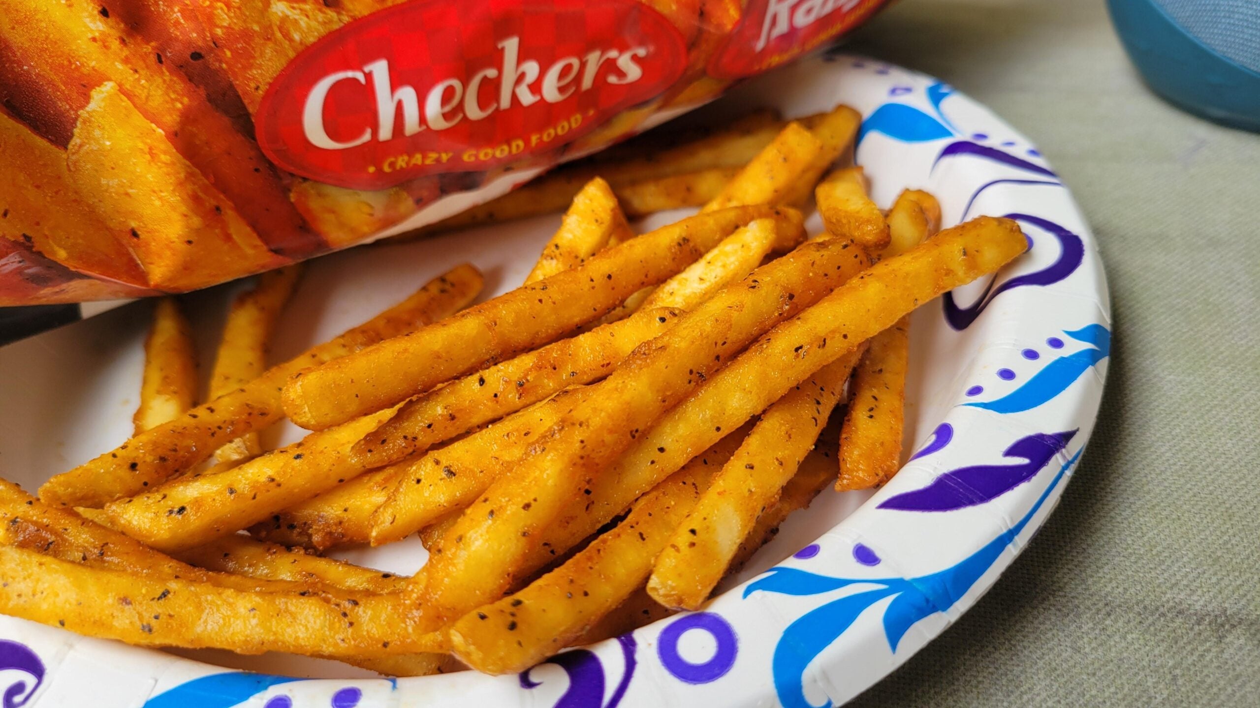 Seasoned French fries from a Checkers bag are served on a white paper plate with blue and purple designs, placed on a light-colored surface. Best fast-food fries