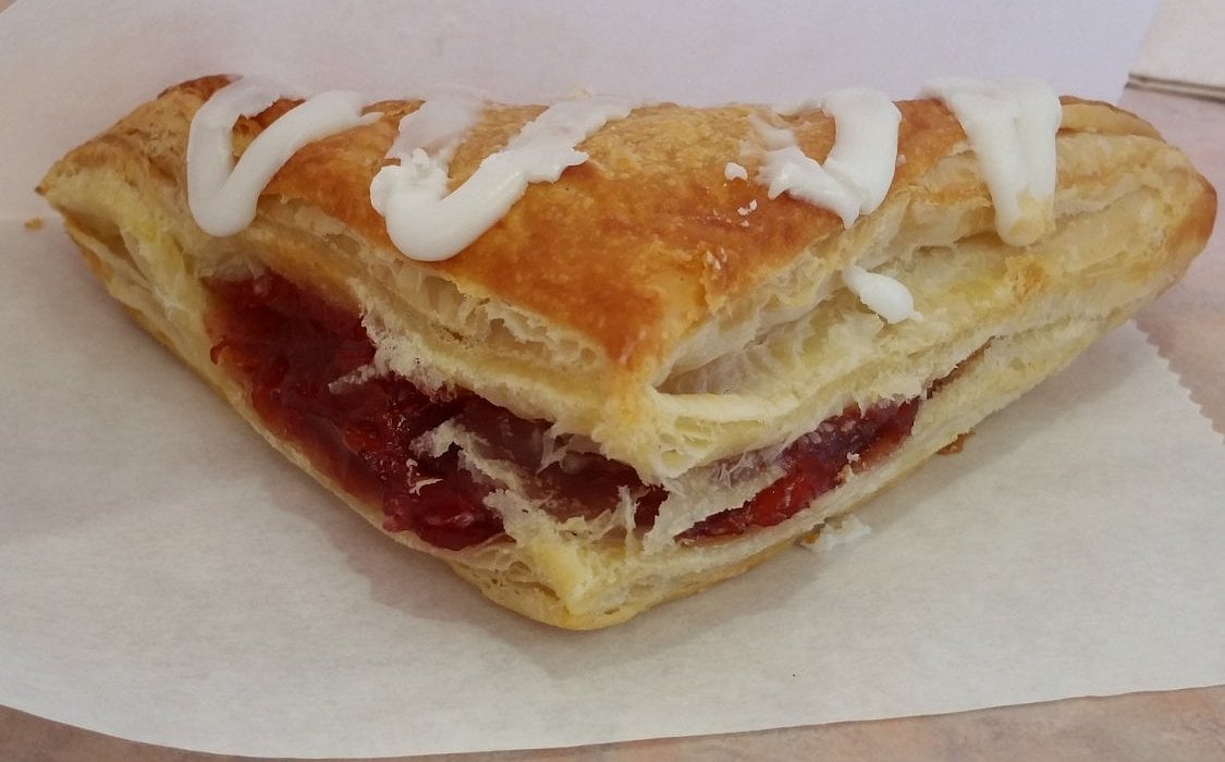 A flaky, golden-brown pastry triangle filled with red fruit jam, topped with a drizzle of white icing, sits on a piece of white parchment paper.