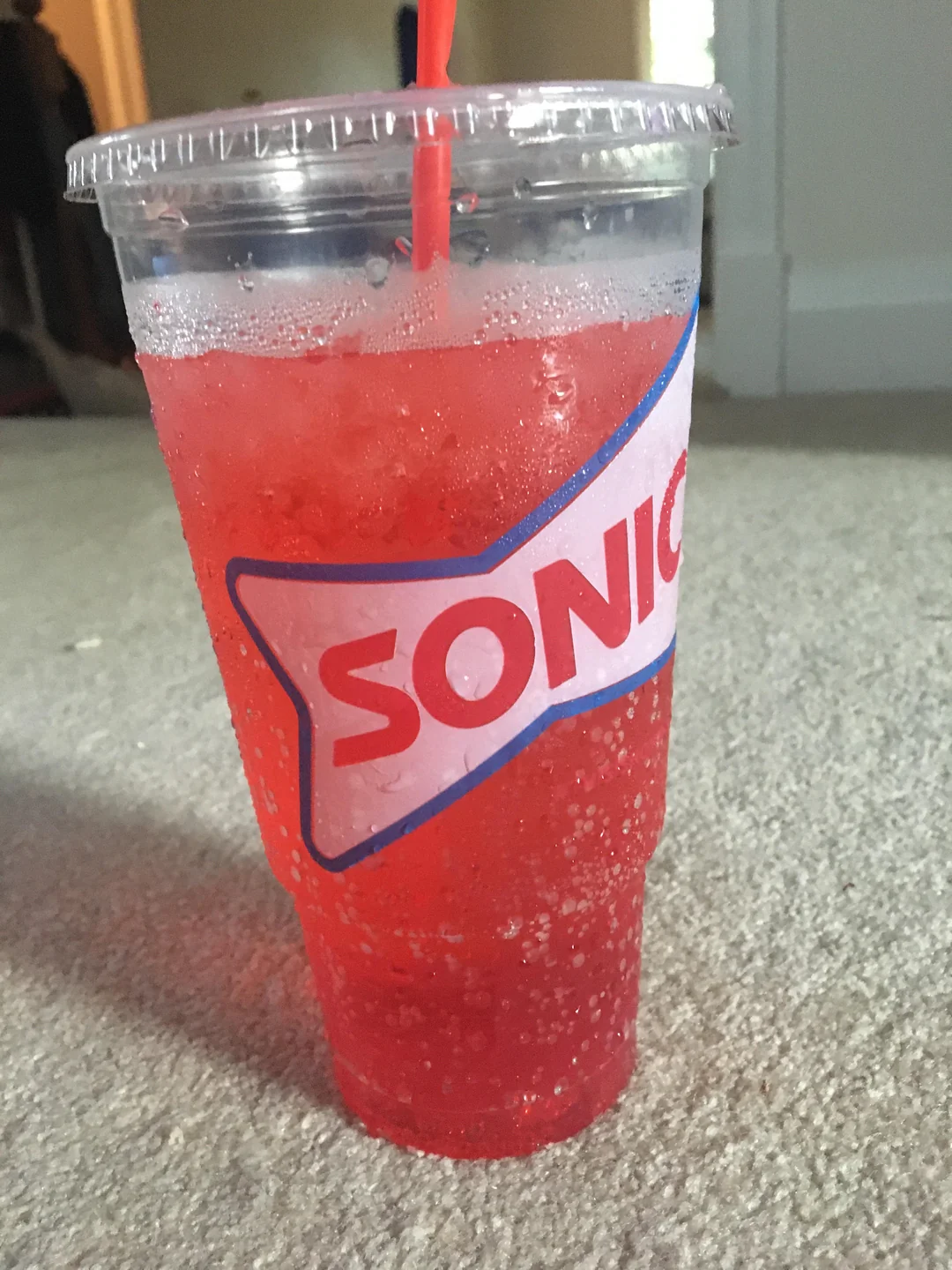 A clear plastic cup filled with a red, bubbly drink and ice from Sonic, with a red straw and condensation on the outside, sits on a light-colored carpeted floor indoors.