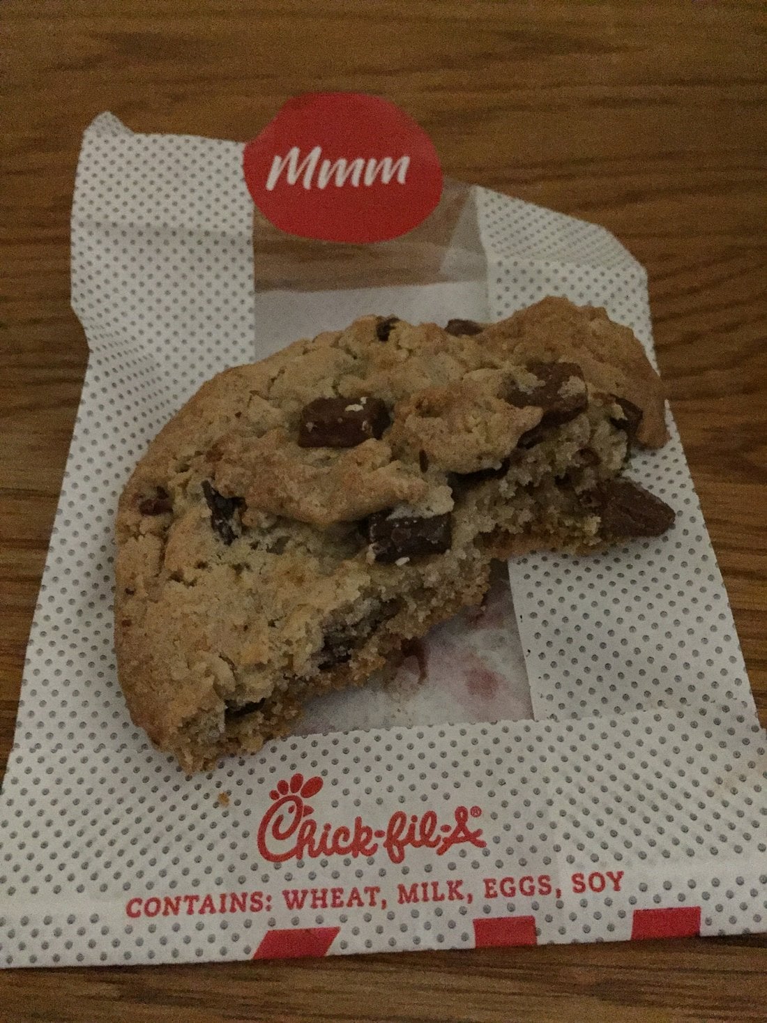 A partially eaten chocolate chip cookie sits on a dotted Chick-fil-A wrapper, which has a red "Mmm" sticker and allergen information noting it contains wheat, milk, eggs, and soy.