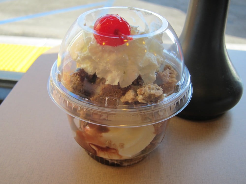 Best fast-food ice cream sundaes. A plastic cup filled with vanilla ice cream, chocolate sauce, crumbled cookies, whipped cream, and topped with a red cherry, covered by a clear dome lid on a table.