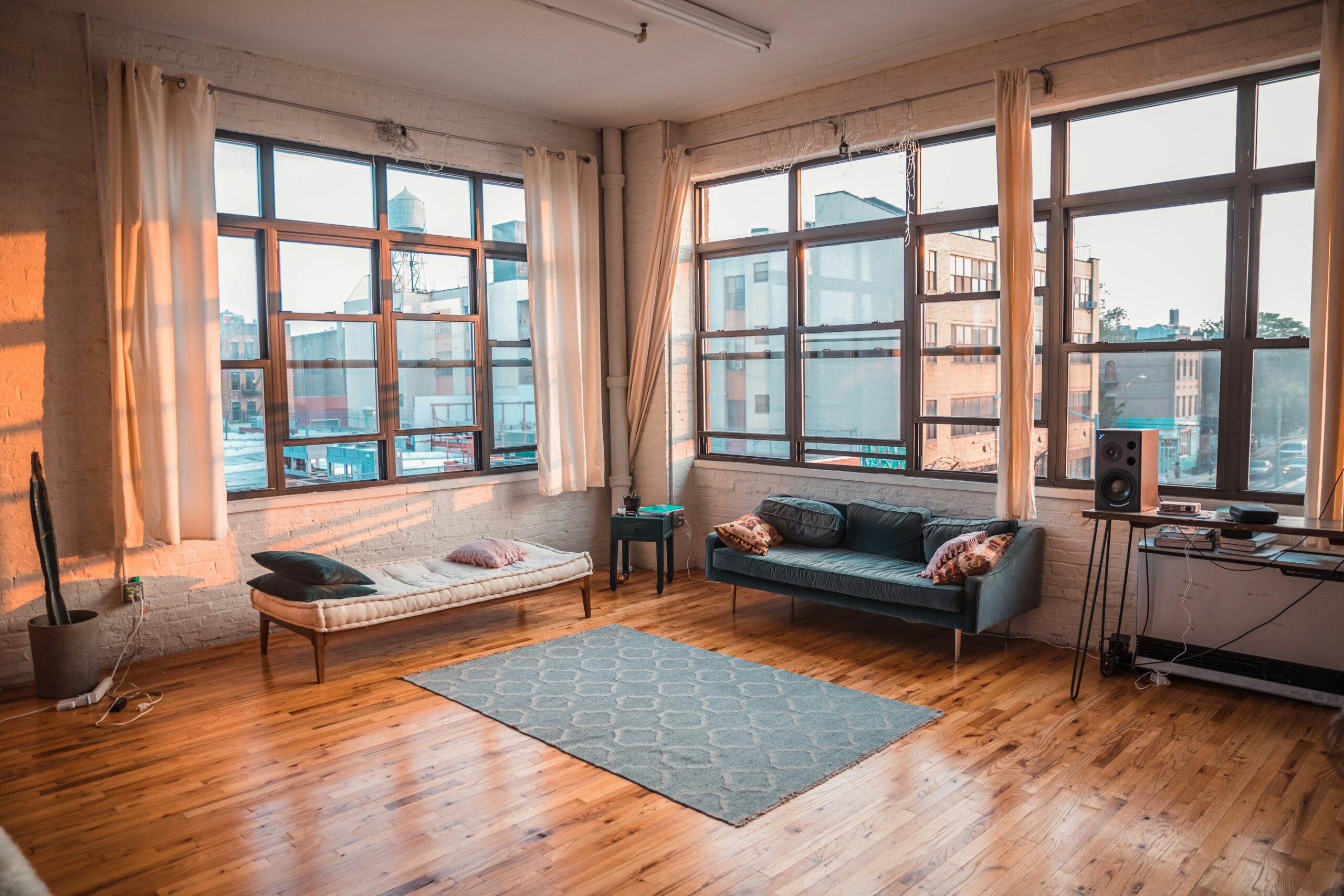 A bright, airy living room with large corner windows, a wooden floor, a patterned rug, a daybed, a blue sofa with pillows, and a small desk with speakers and electronics near the windows.