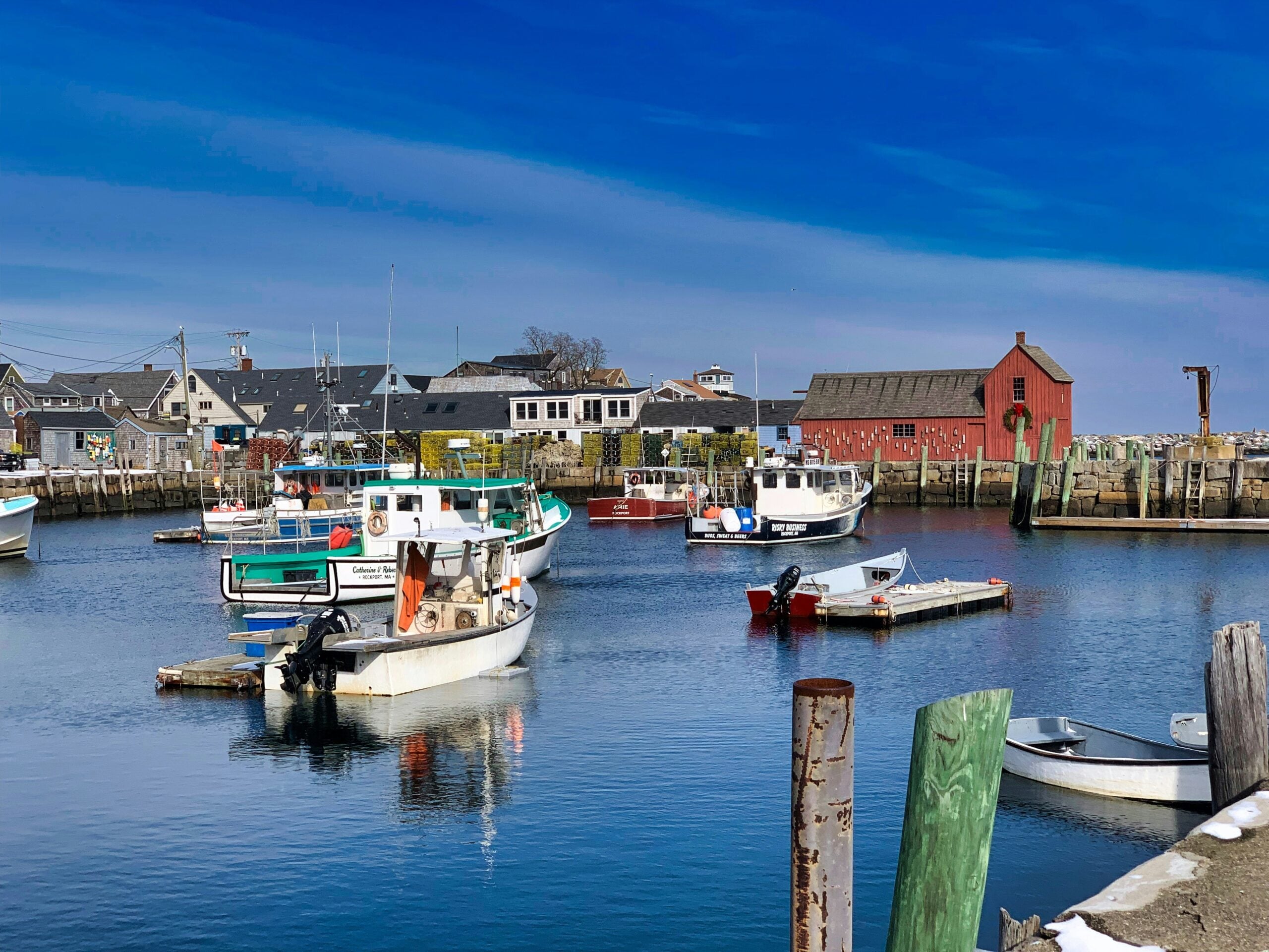 Colorful fishing boats are docked in a small harbor with wooden piers, buildings, and a prominent red shack under a bright blue sky. The water is calm, reflecting the boats and surrounding structures.