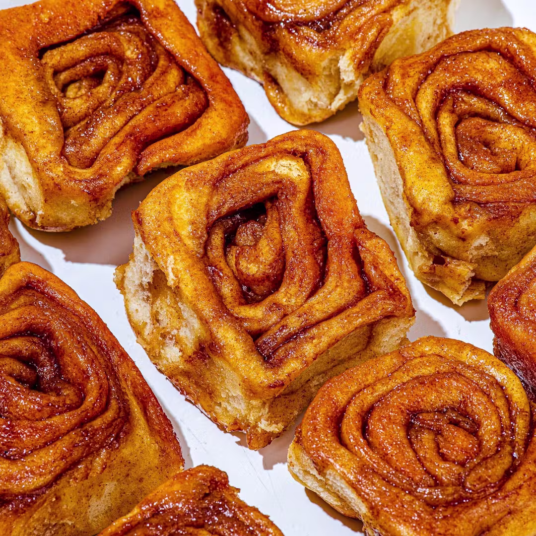 Close-up of several golden-brown cinnamon rolls with swirls of cinnamon and sugar, arranged on a white surface. The rolls look soft, fluffy, and slightly glossy, suggesting they are freshly baked.