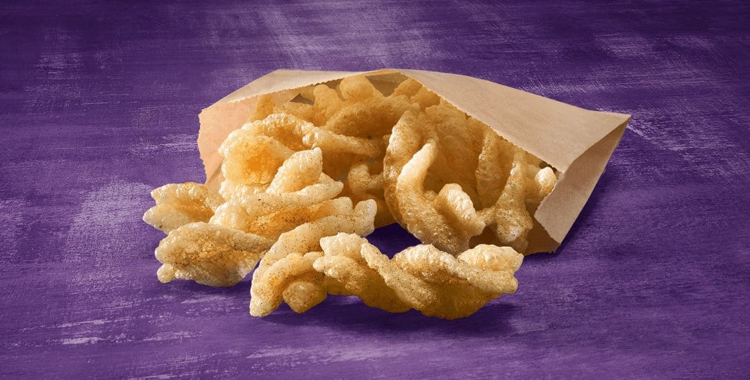 A brown paper bag tipped over on a purple surface, spilling out several golden, crispy pork rinds—an unexpected crunch among classic fast-food desserts.