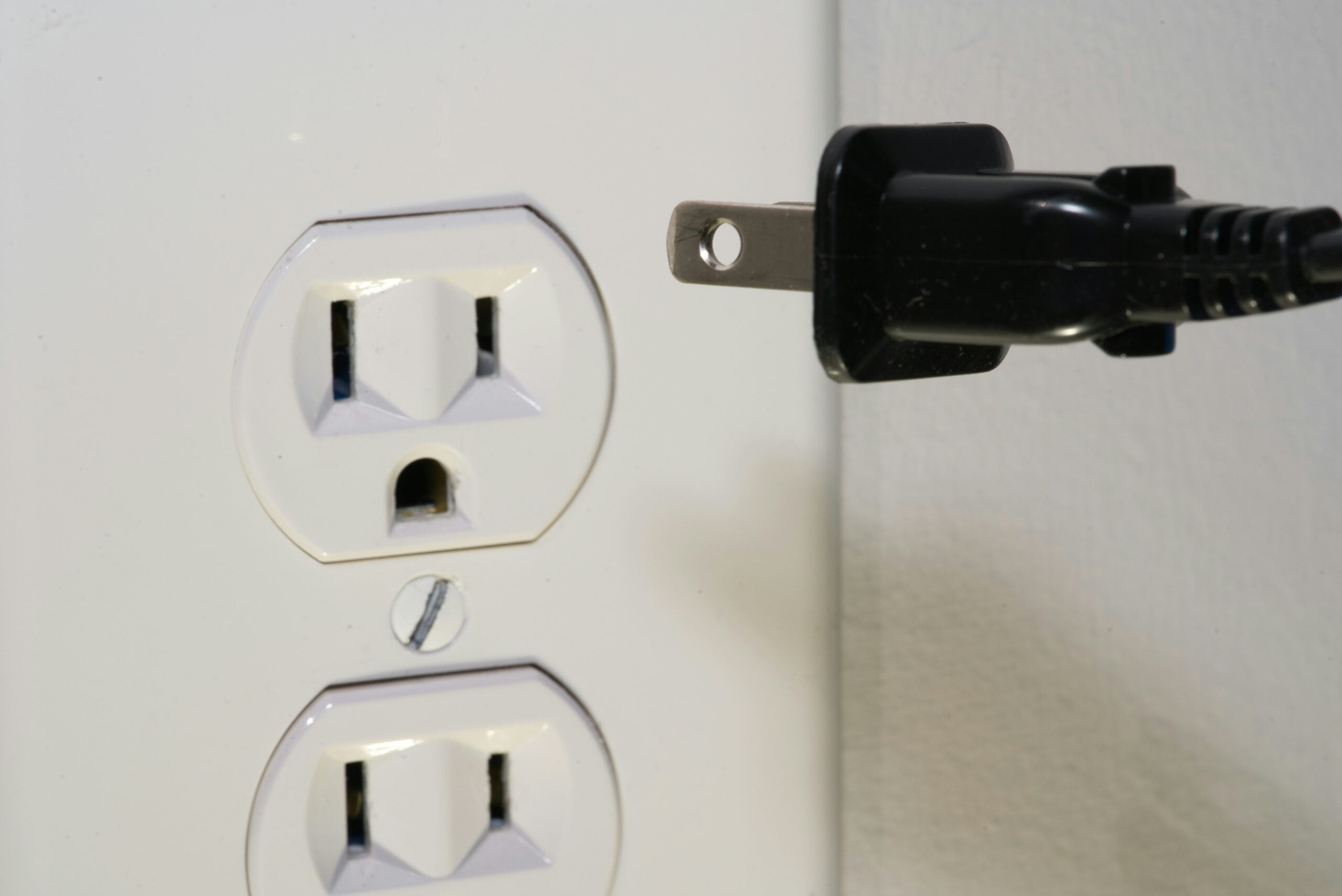 A close-up of a black electrical plug being held near a white wall power outlet, ready to be plugged in. The outlet has two sockets, and the background is a plain wall.