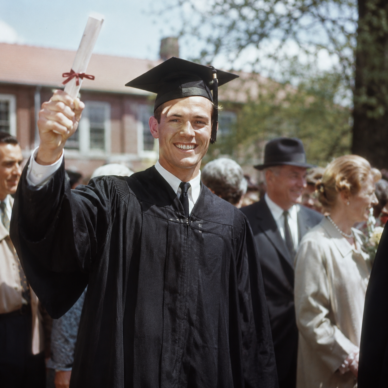A young man in a graduation cap and gown smiles and holds up his diploma, standing outdoors among a crowd of people during a graduation ceremony in the early 1960s on a single income.