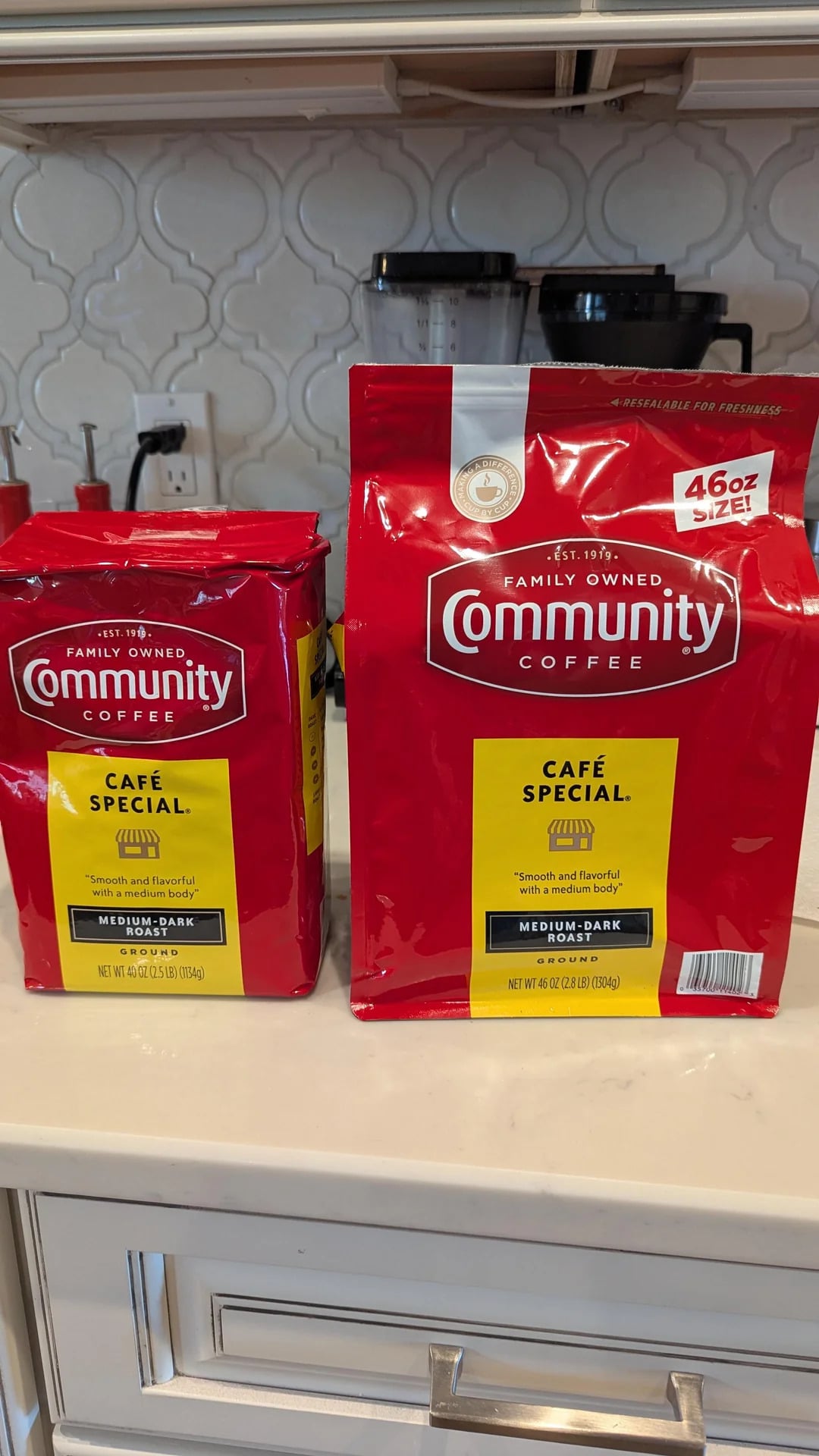Two red bags of Community Coffee Café Special Medium-Dark Roast, a standout among coffee brands, sit on a kitchen counter—one 32-ounce and one 46-ounce. A coffee maker is visible in the background.