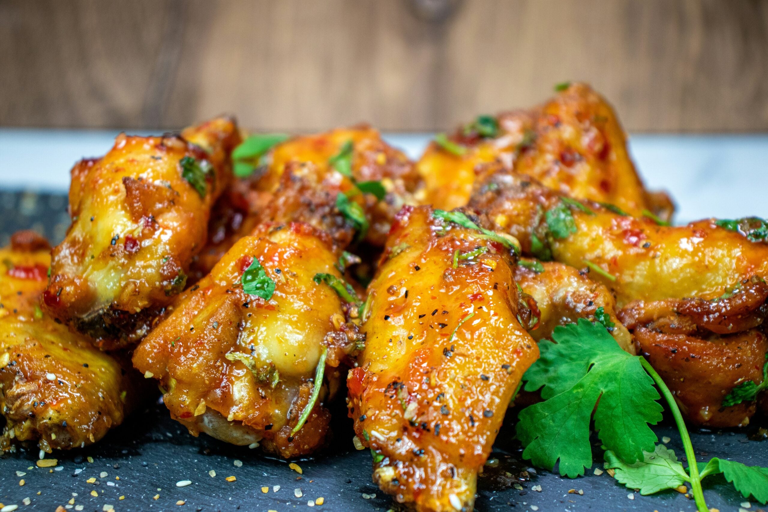 A close-up of several spicy baked chicken wings garnished with herbs and cilantro, resting on a dark slate surface with a wooden background.