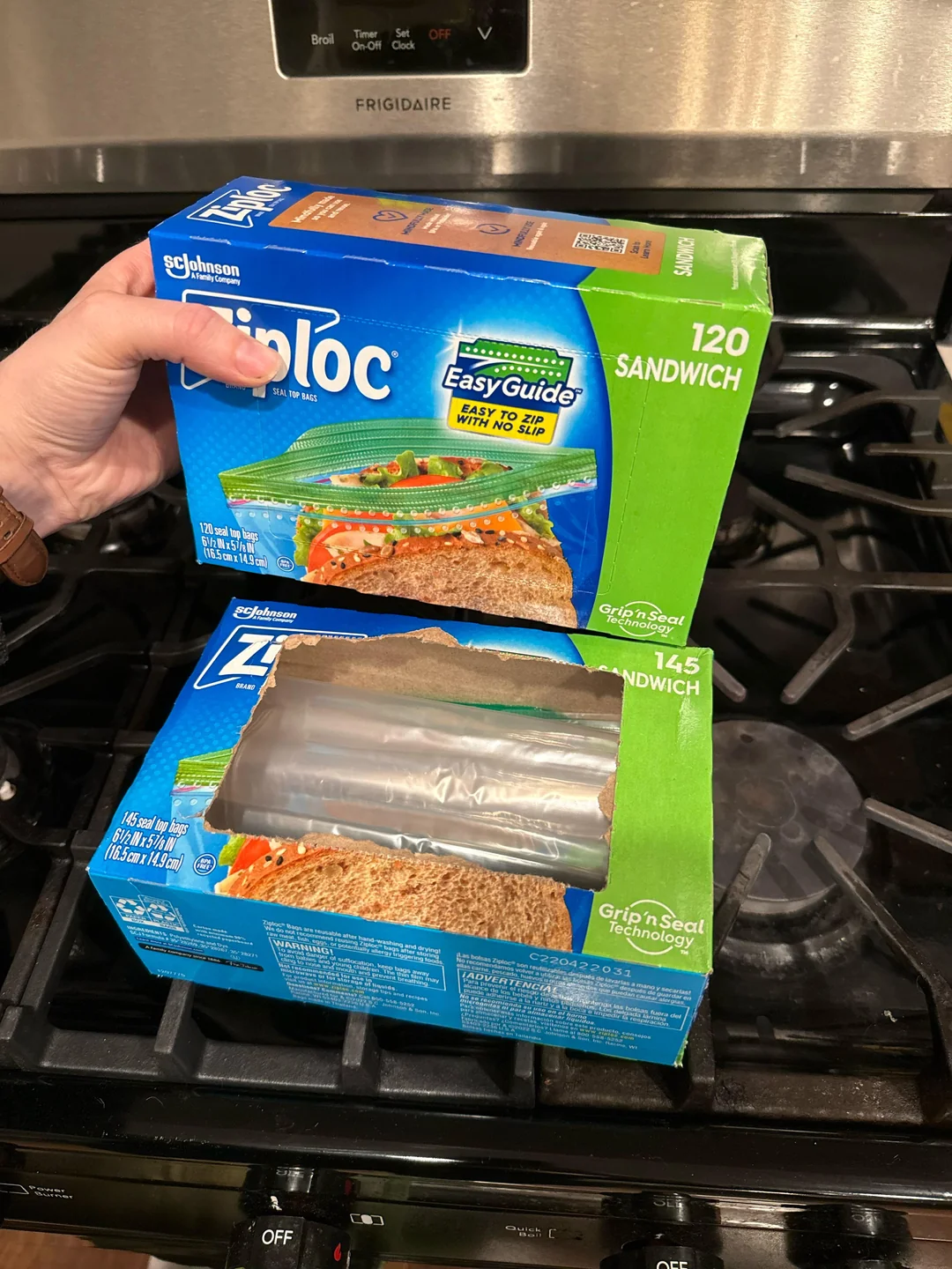 A person holds a box of Ziploc sandwich bags above another open box of Ziploc sandwich bags on a stove. Both boxes are blue and green, with plastic bags visible in the open box.