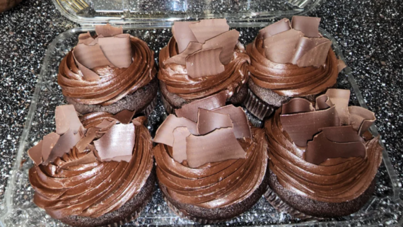 Six chocolate cupcakes with swirled chocolate frosting are topped with large, curly chocolate shavings&mdash;a decadent addition to your favorite Costco Bakery Items&mdash;arranged in a clear plastic container on a dark, speckled countertop.