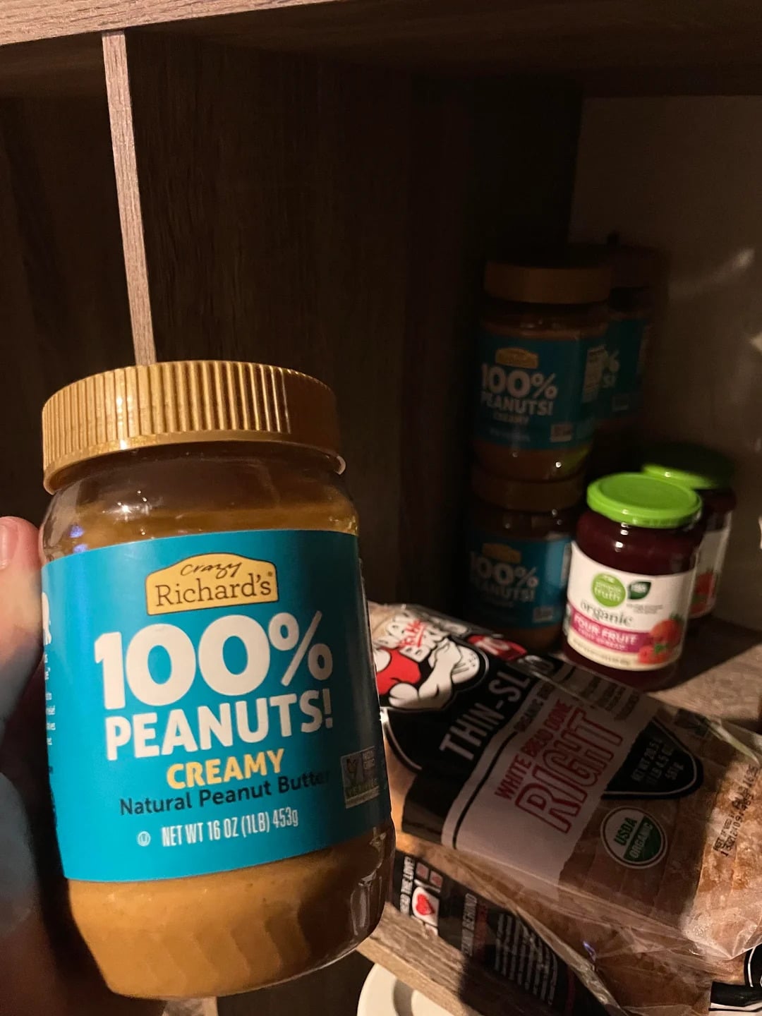 A hand holds a jar of Crazy Richard's 100% Peanuts! Creamy natural peanut butter—one of the best peanut butter brands—in front of a pantry shelf stocked with more jars, a loaf of bread, and strawberry fruit spread.