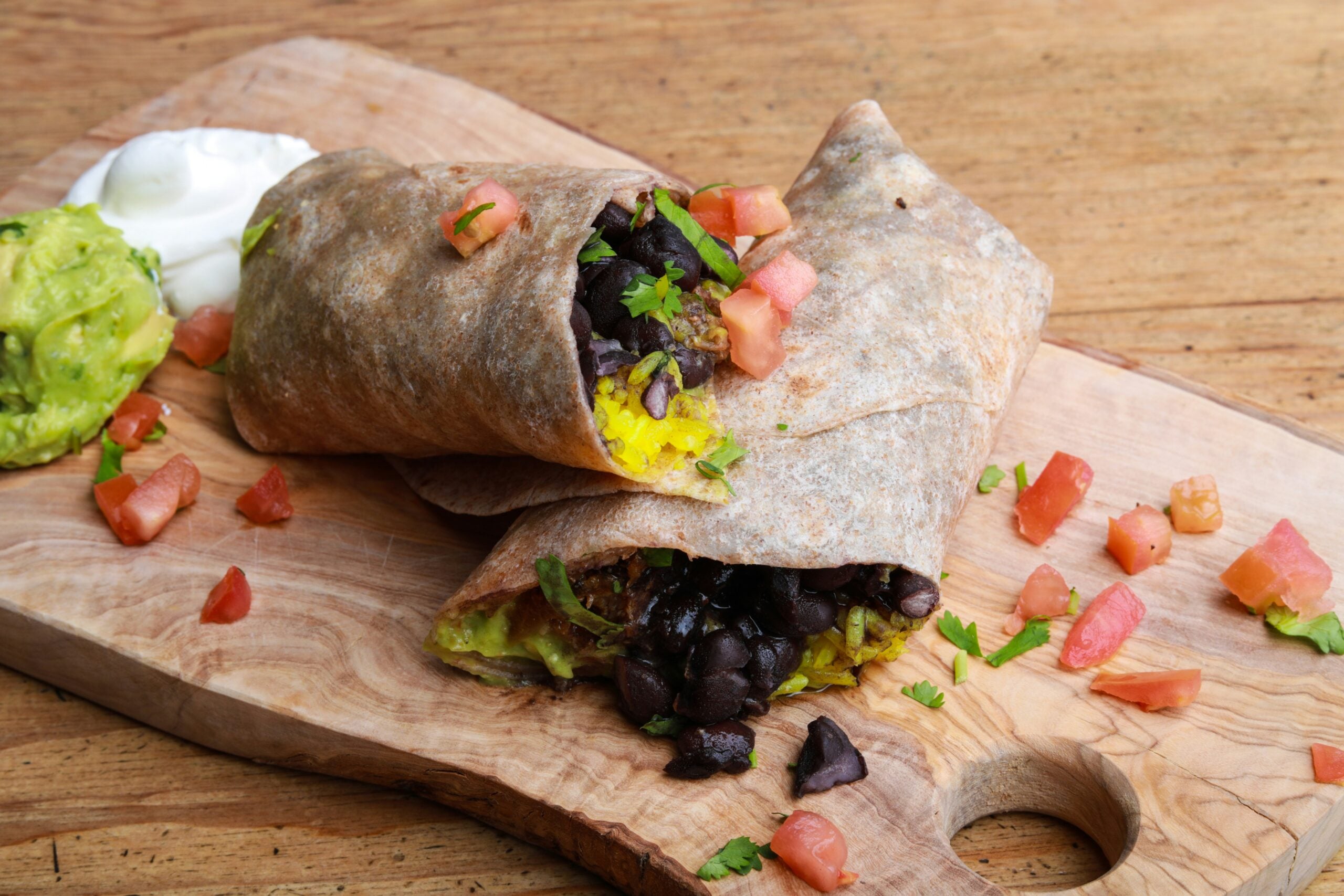 A whole wheat burrito cut in half on a wooden board, filled with black beans, yellow rice, guacamole, and topped with diced tomatoes and cilantro. Sour cream and more guacamole are on the side.
