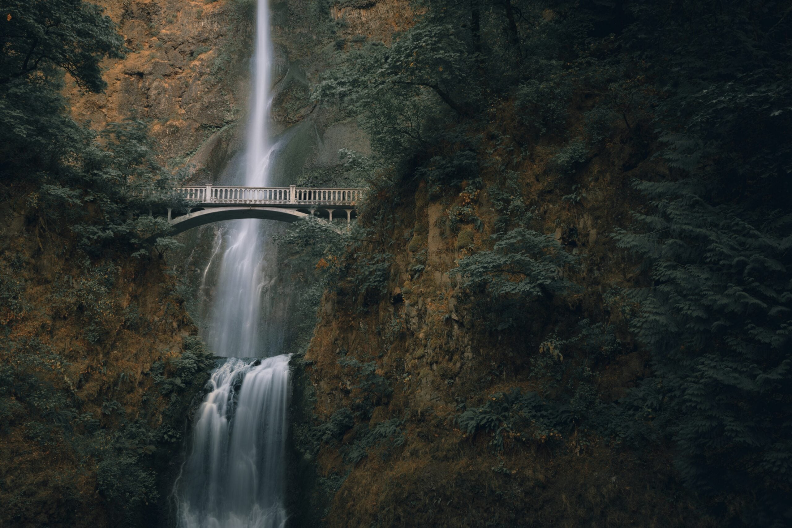 A tall waterfall cascades down a rocky cliff, with a scenic arched footbridge crossing in front of the falls, surrounded by dense green foliage.