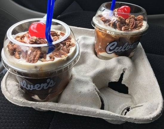 Two Culver’s sundaes topped with pecans, a cherry, and blue spoons sit in a gray cardboard drink holder on a car seat. The sundaes have visible layers of vanilla ice cream and chocolate sauce.