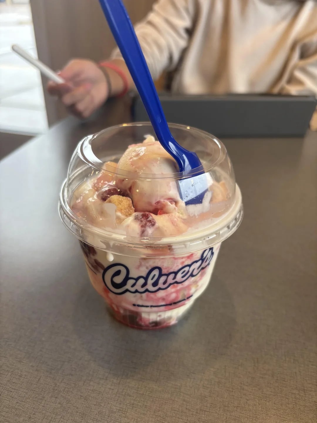 A Culver’s frozen custard dessert, one of the tastiest fast-food desserts, features strawberries and shortcake pieces in a clear cup with a blue spoon. In the blurred background, someone uses a tablet and phone at the table.