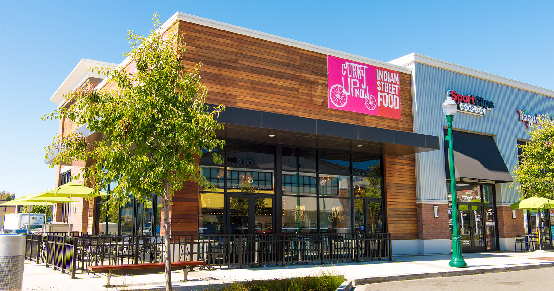 A modern restaurant with wood and brick exterior, large windows, and patio seating sits under a clear blue sky. A pink sign reads "Curry Up Now: Indian Street Food." There are green umbrellas and a nearby tree. Up-and-coming fast-food chains.