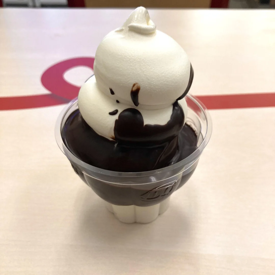 A clear plastic cup filled with vanilla soft serve ice cream, topped with thick chocolate sauce, sits on a light-colored table with a red design in the background.