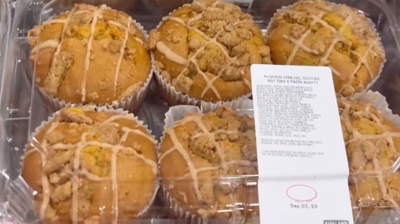 A clear plastic container holds six pumpkin streusel muffins topped with crumbly streusel and icing drizzle. A white label with product information and an expiration date is attached to the container.