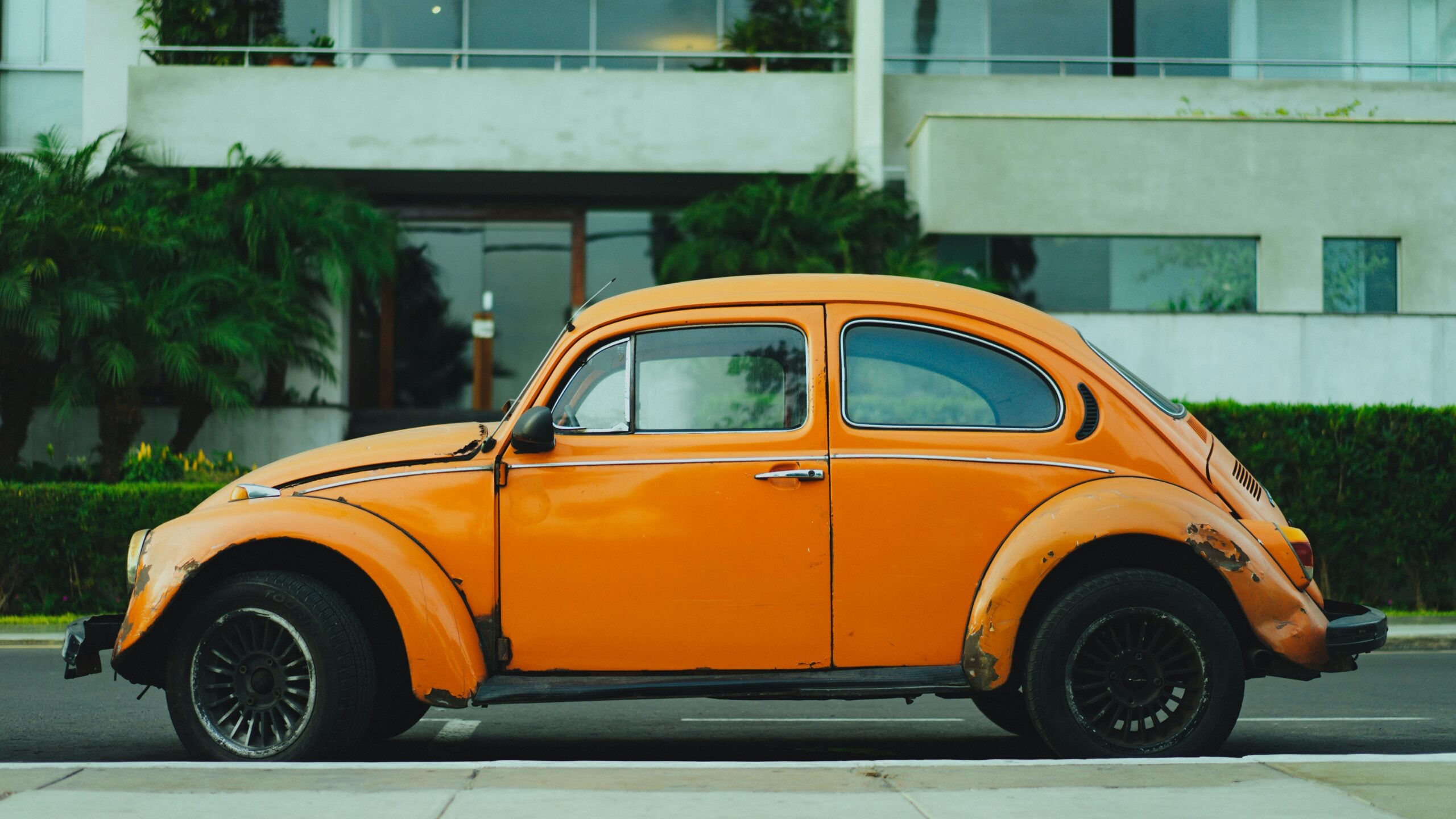 An old, orange Volkswagen Beetle with visible rust and wear is parked on the street in front of a modern building with greenery and large windows.