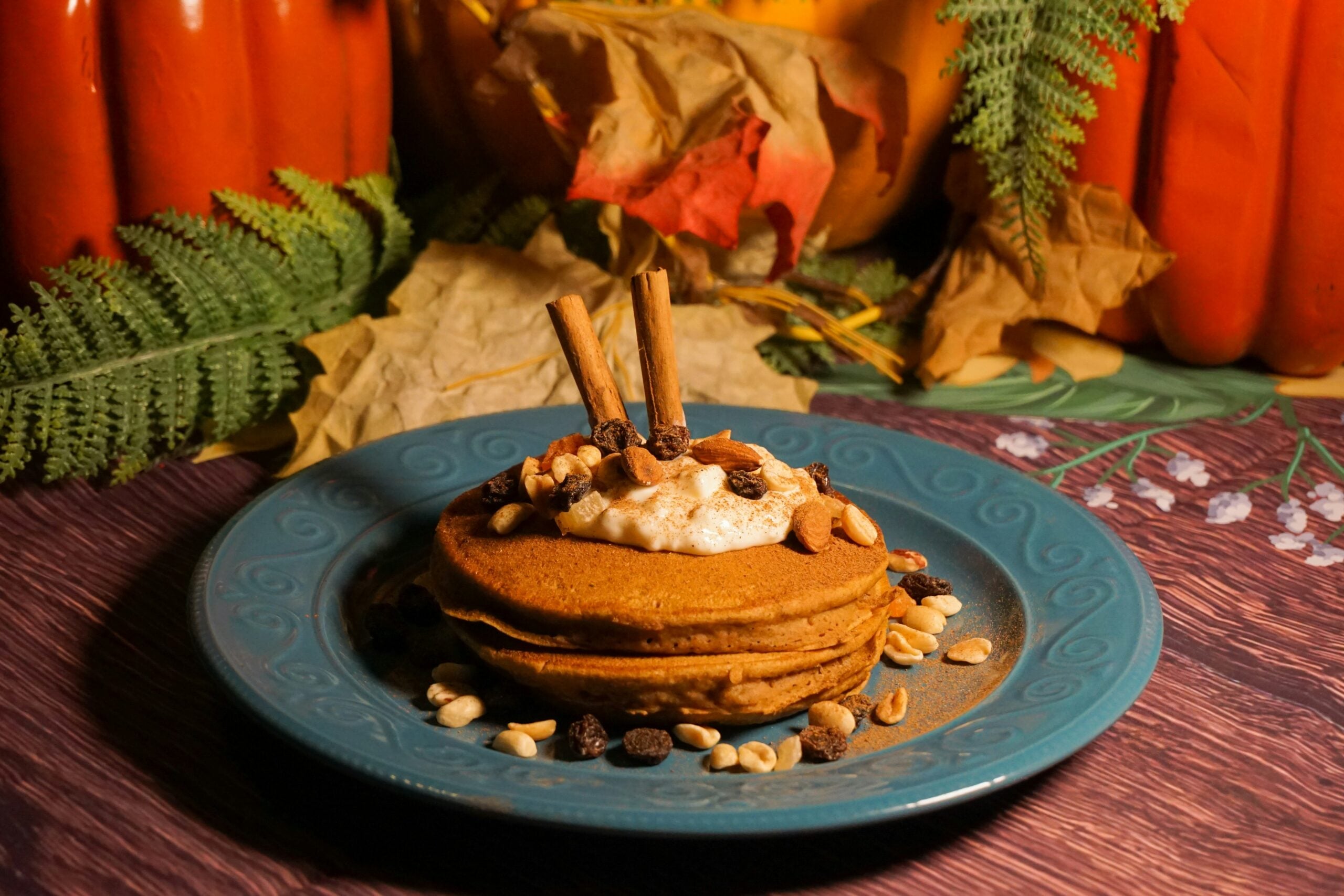 A stack of pancakes topped with whipped cream, cinnamon sticks, nuts, and raisins sits on a blue plate. Autumn leaves, ferns, and pumpkins decorate the background, creating a cozy fall atmosphere.