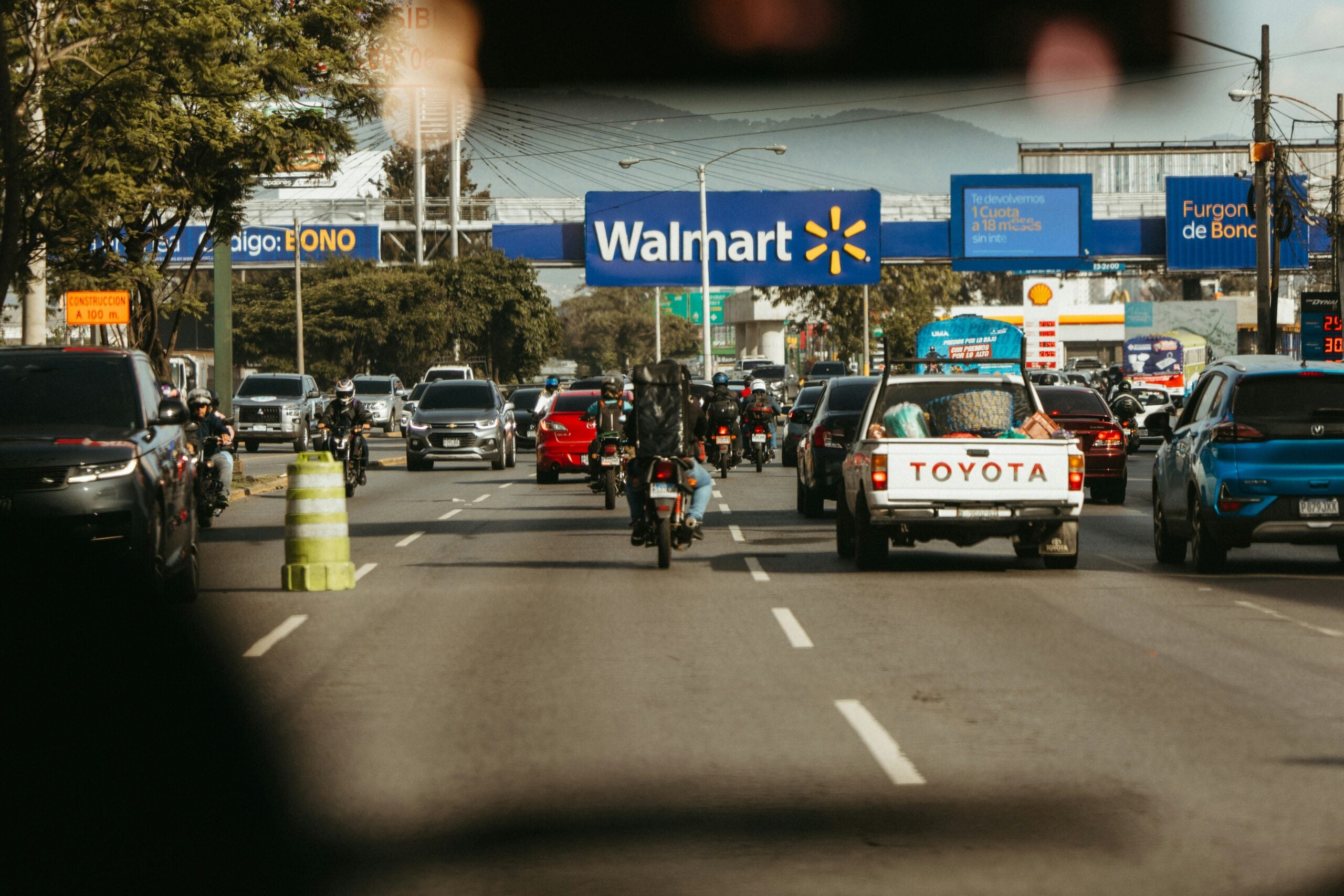 Busy city street with several cars, motorcycles, and a white Toyota pickup truck heading towards a Walmart store sign on a sunny day. Trees and various business signs are visible along the road.