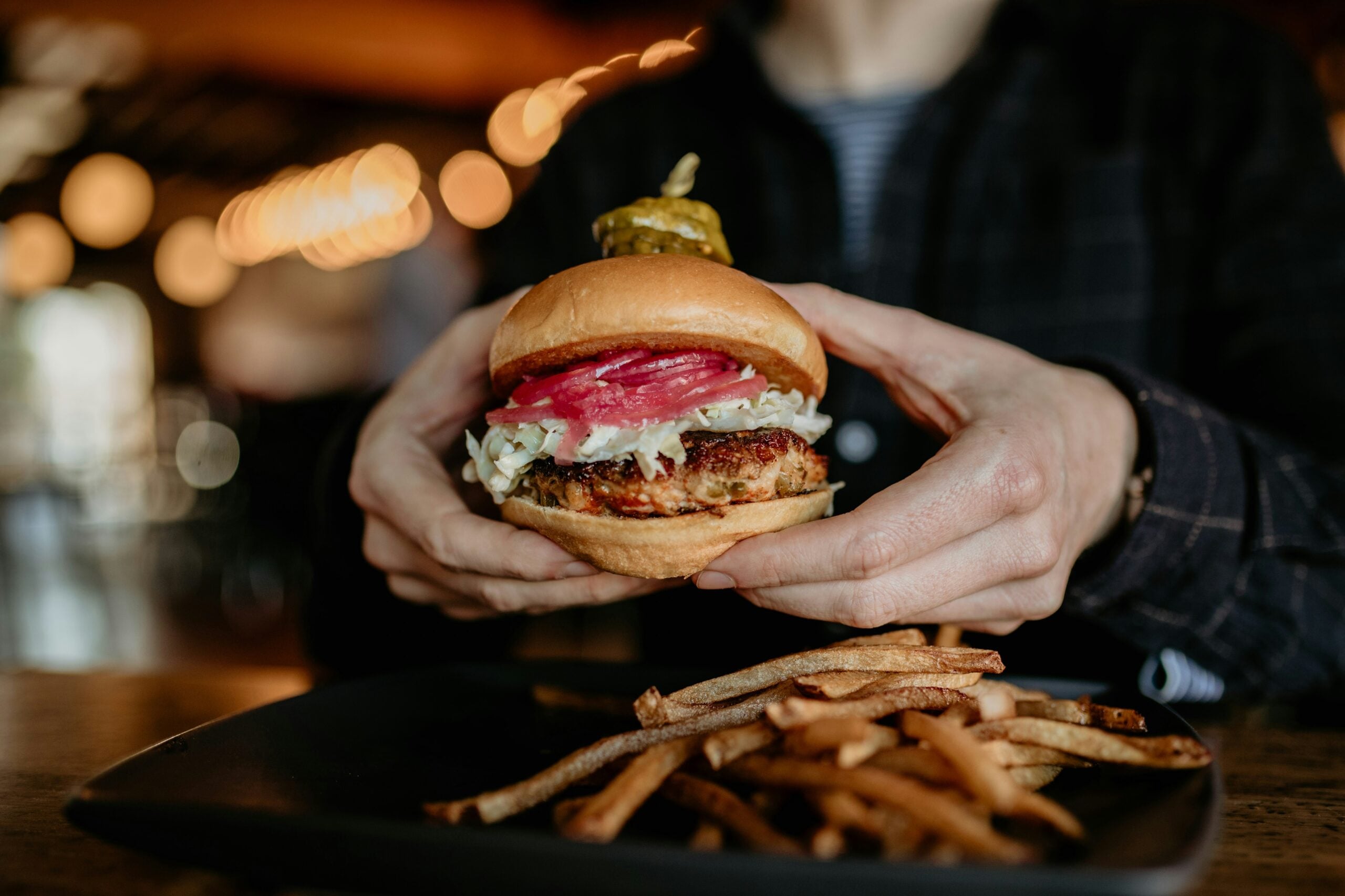 A person holding a burger with lettuce, pickled onions, and a pickle on top, with a plate of thin French fries on the table in front of them. The background is softly blurred.