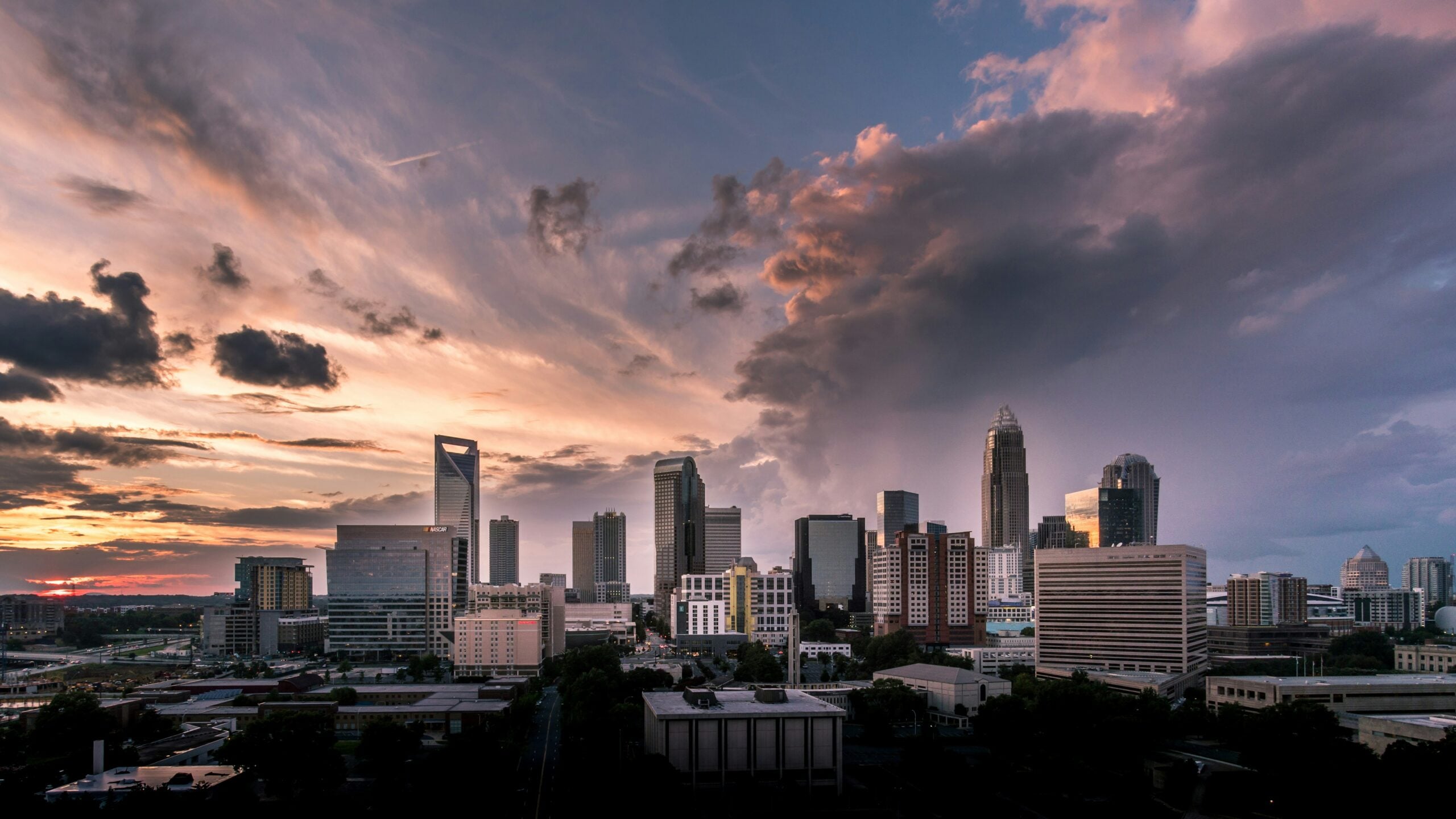 A city skyline at sunset with tall modern buildings and dramatic clouds in the sky, casting a warm and dusky light over the urban landscape.