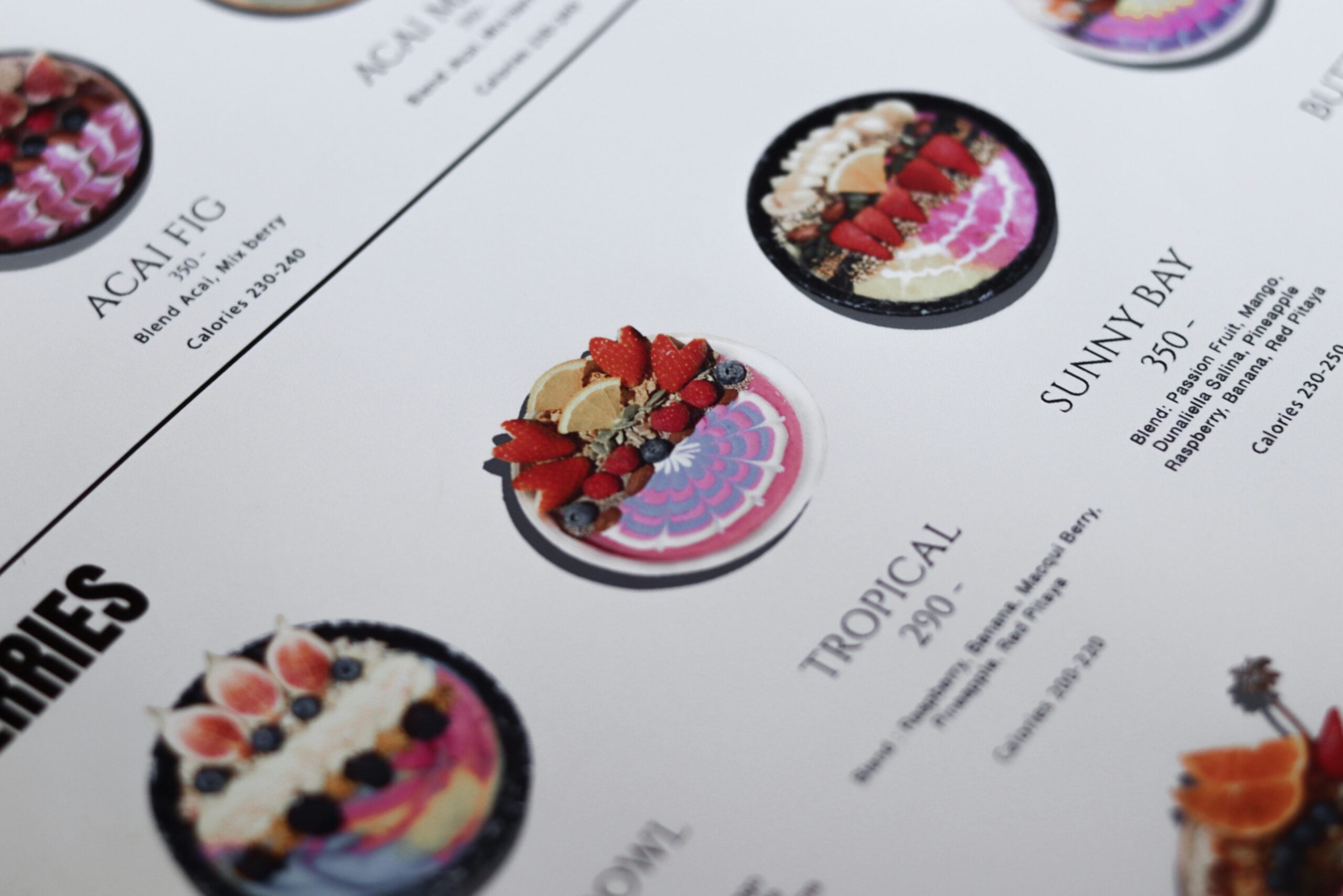 Close-up of a menu featuring colorful smoothie bowls with artistic fruit toppings. The bowls are labeled with names like "Tropical" and "Sunny Bay," and descriptions, prices, and calorie counts are visible beneath each item.