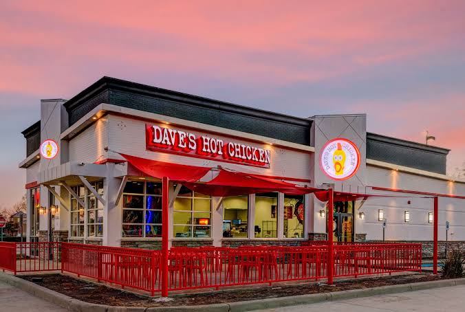 A Dave’s Hot Chicken restaurant with red accents, outdoor seating, and large illuminated signs, photographed at sunset under a pink and blue sky.