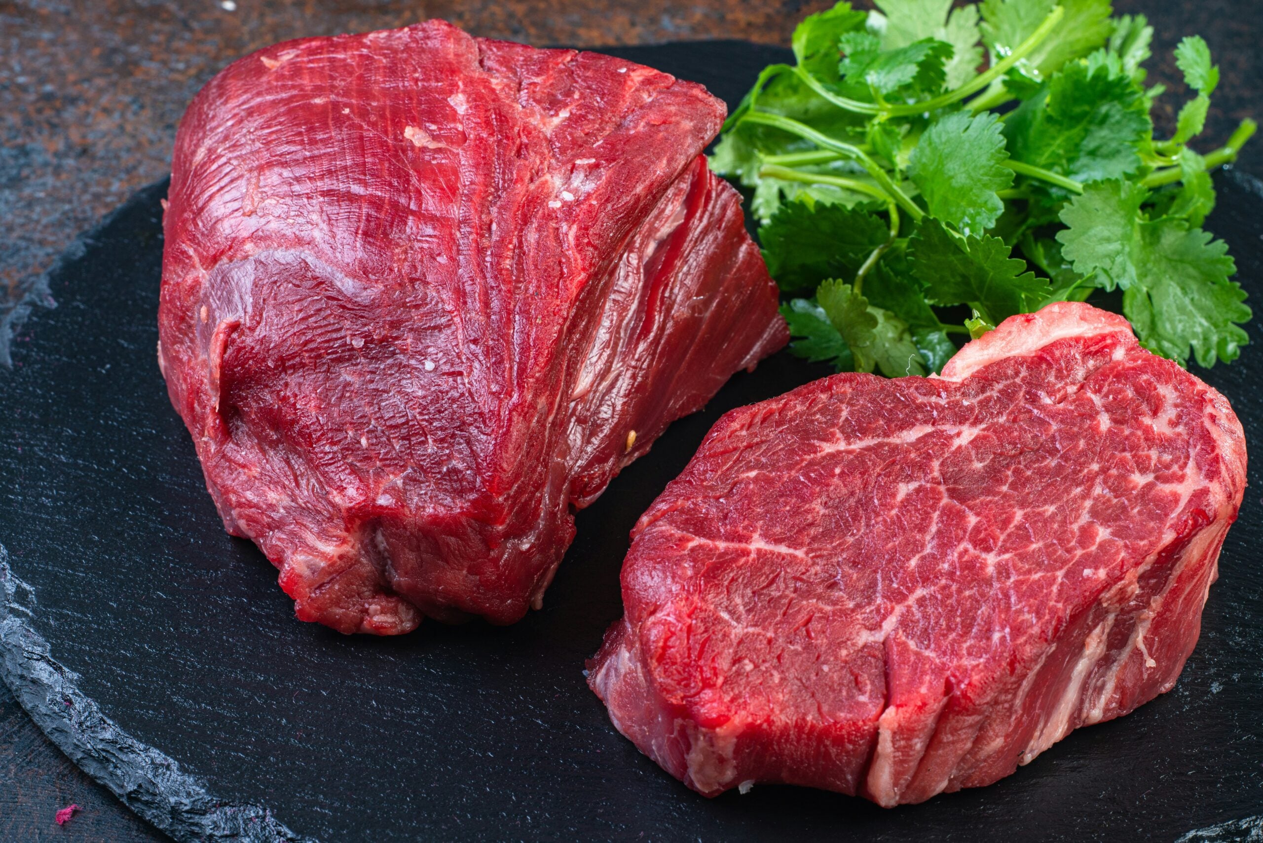 Two raw beef steaks are placed on a round slate board with a bunch of fresh cilantro leaves beside them. The meat is red and marbled, ready for cooking.
