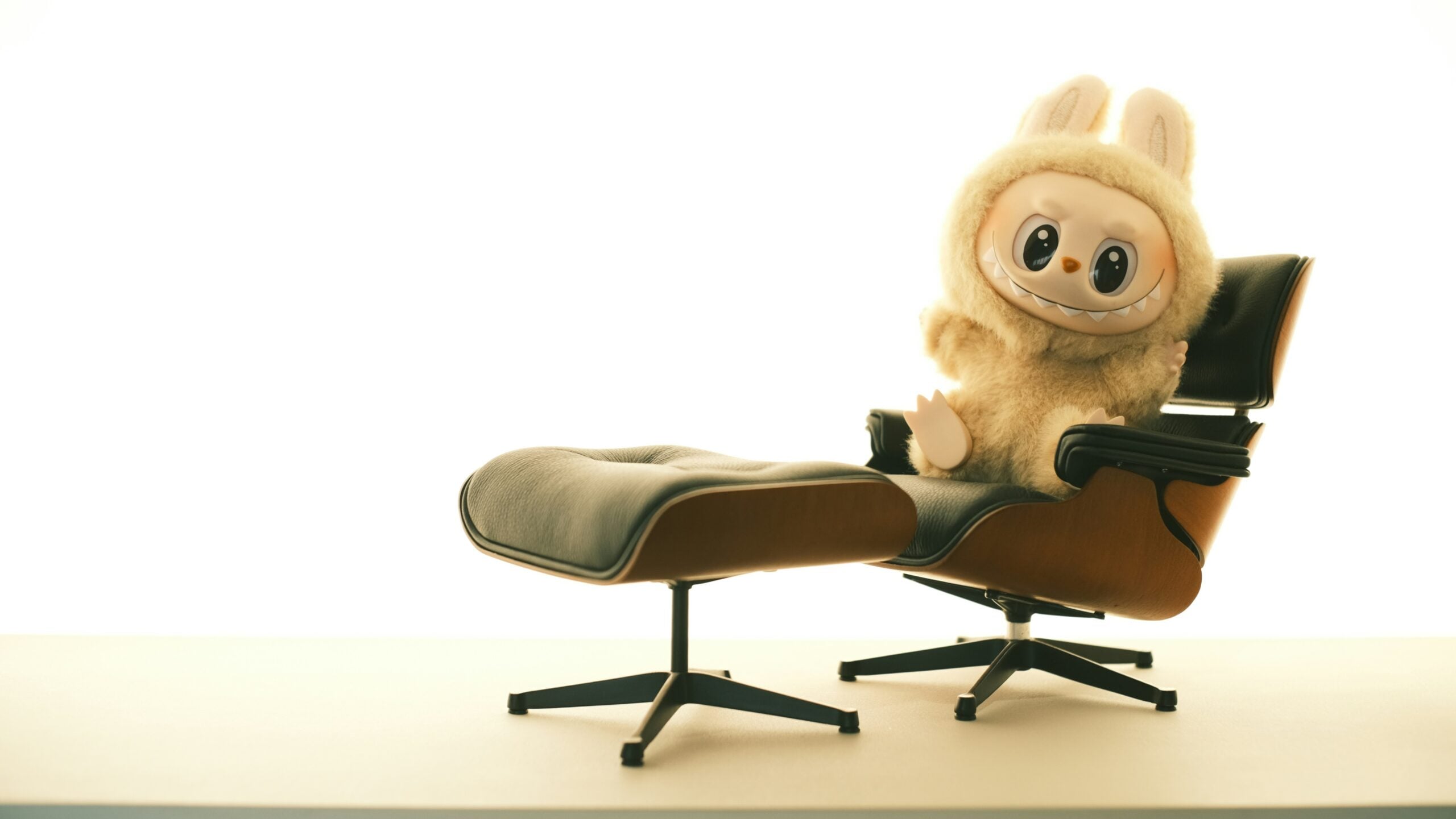 A cute plush toy with big eyes, wearing a fluffy bunny costume, sits on a stylish lounge chair with an attached ottoman, against a bright, minimalist background.