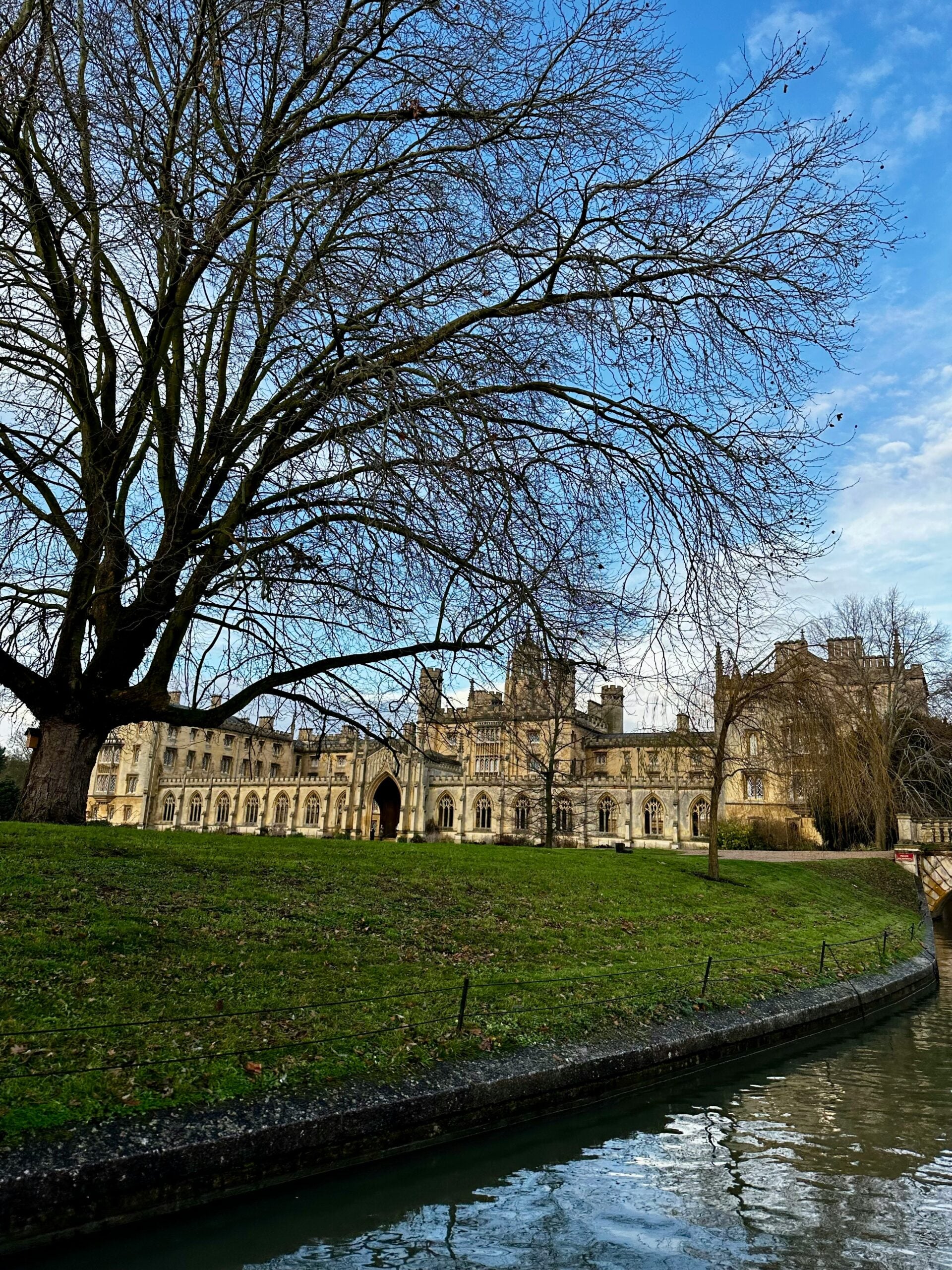 A historic stone building stands behind a large leafless tree on a grassy lawn, with a canal curving along the edge under a blue sky with scattered clouds.