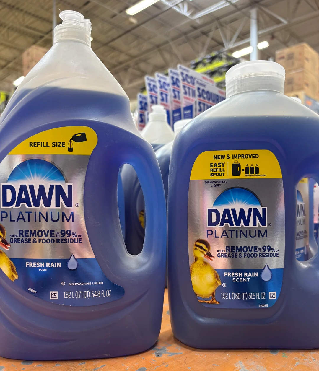 Two large bottles of Dawn Platinum dish soap with "Fresh Rain" scent are displayed on a store shelf. The labels highlight "Removes 99% grease & food residue" and feature images of a duckling.