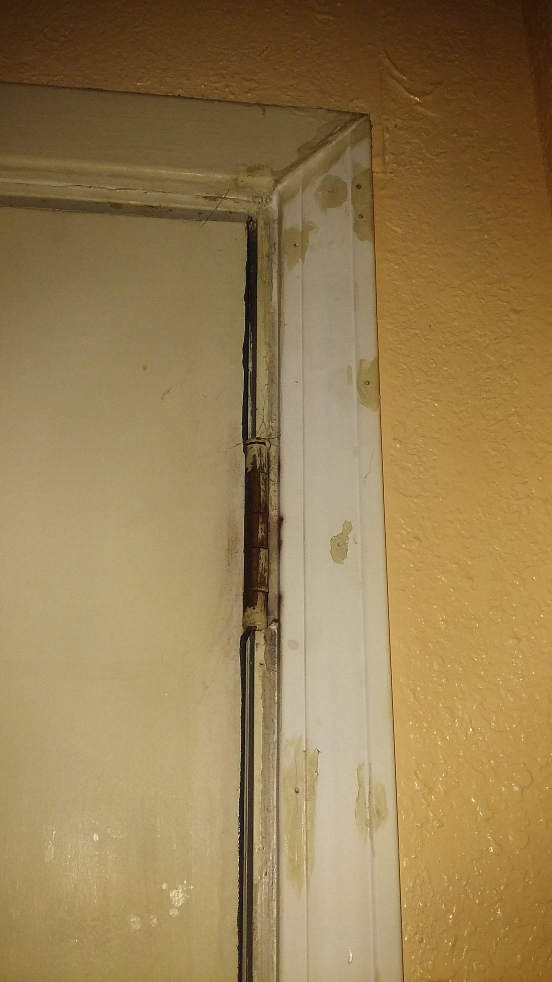 A close-up of a white door hinge with visible rust and paint chipping on the door frame, against a textured tan wall—a detail from properties potentially slated for renovation by major Hotel Brands 2026. Multiple patched areas are seen on the trim.